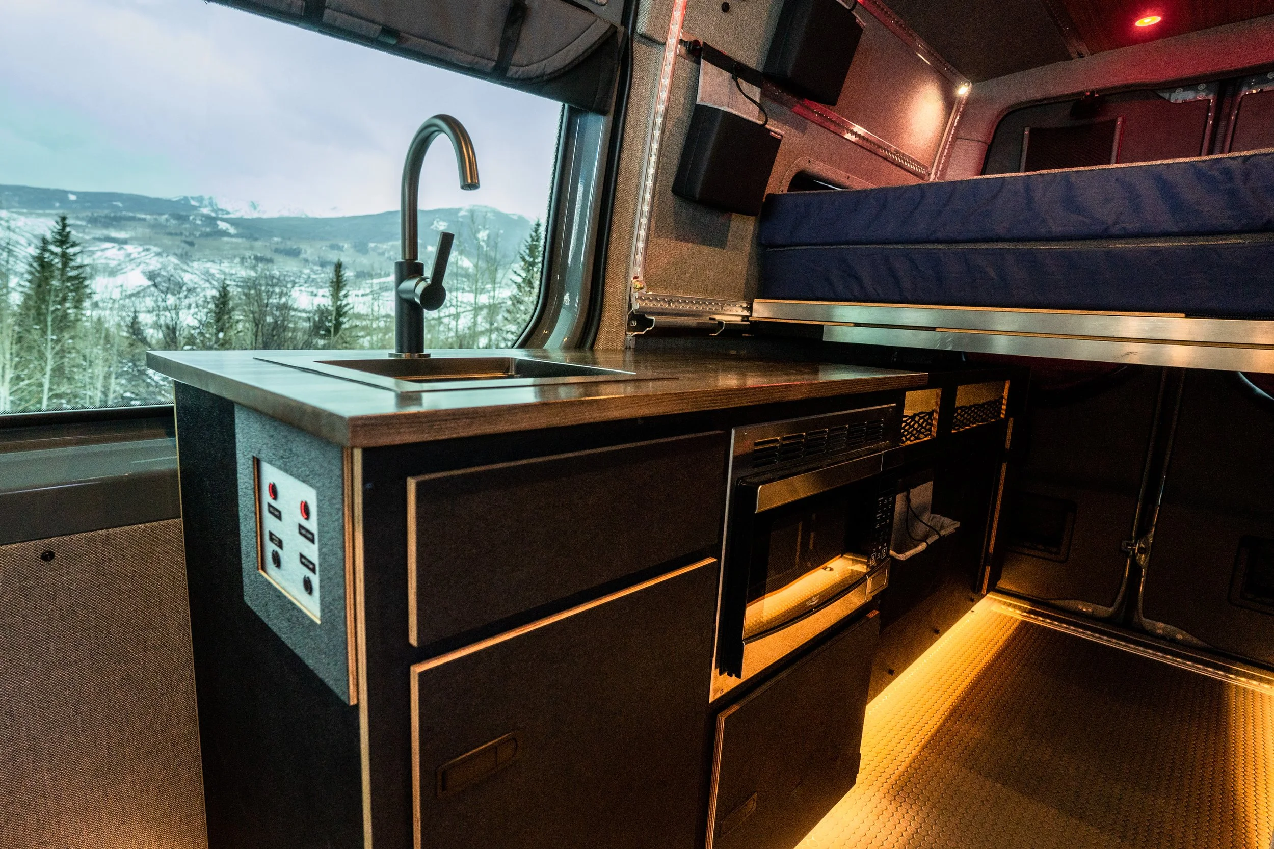 Interior of a converted van showing a small kitchen area with a countertop sink, oven, and a sleeping platform above, with a mountain and forest view outside the window.