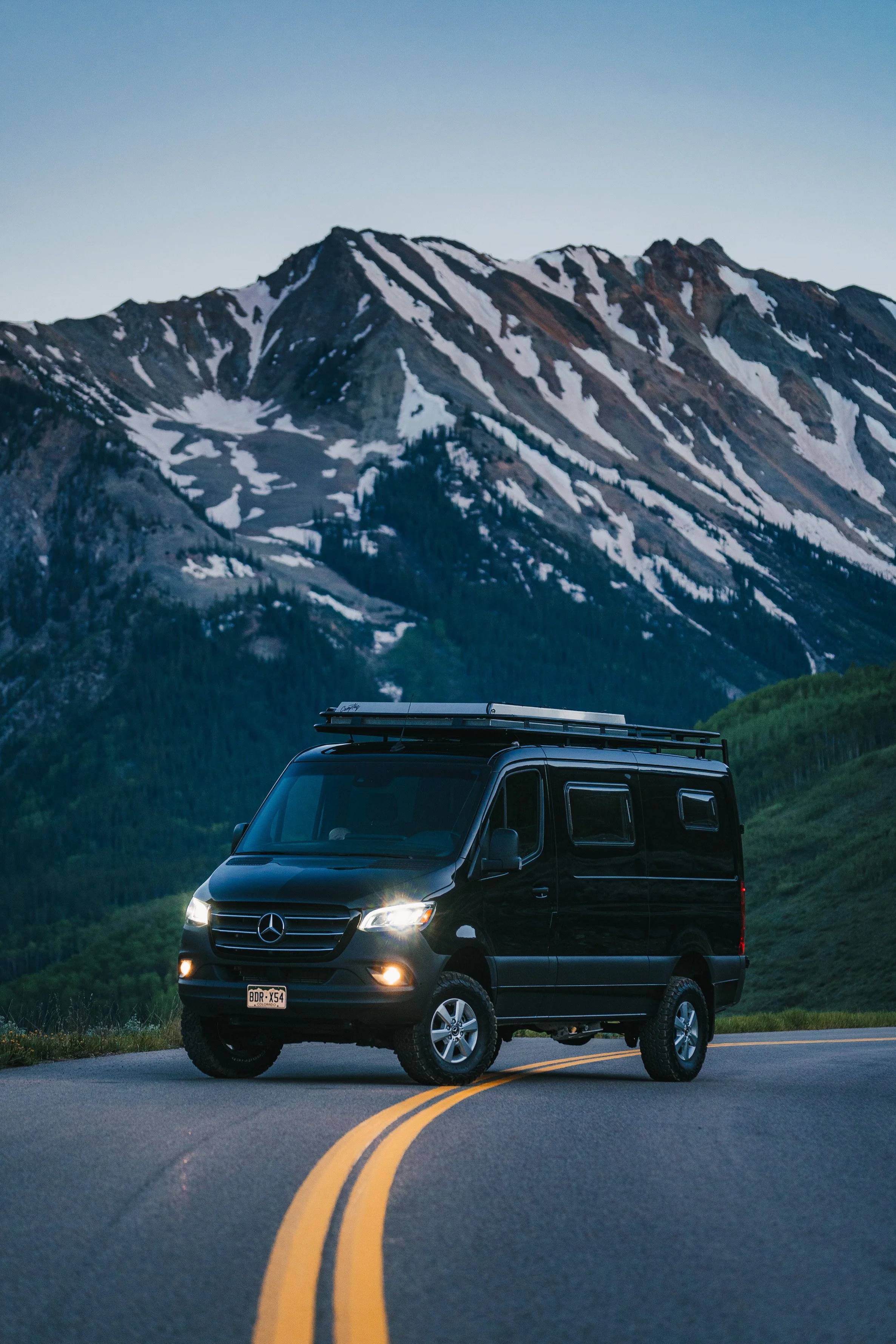 A black camper van parked on a winding mountain road with snow-capped peaks in the background during dusk.