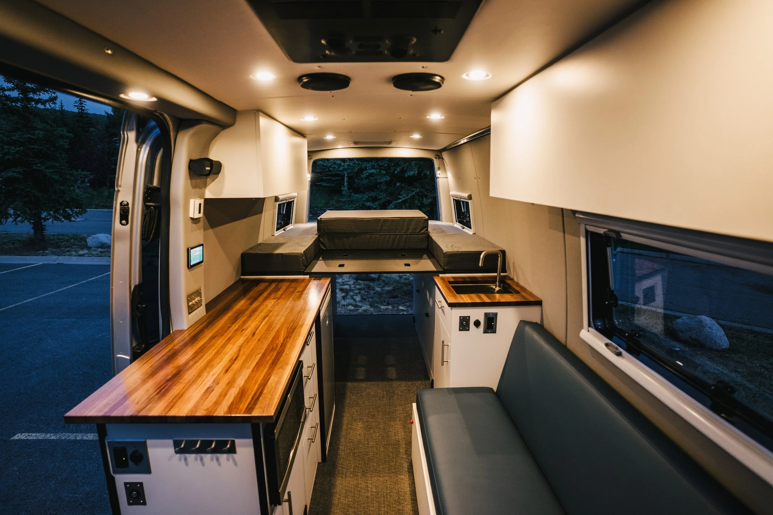 Interior of a converted van or small camper with a wooden countertop, a small sink, a gray bench seat, and a sleeping area at the back with cushions, illuminated by ceiling lights at dusk.