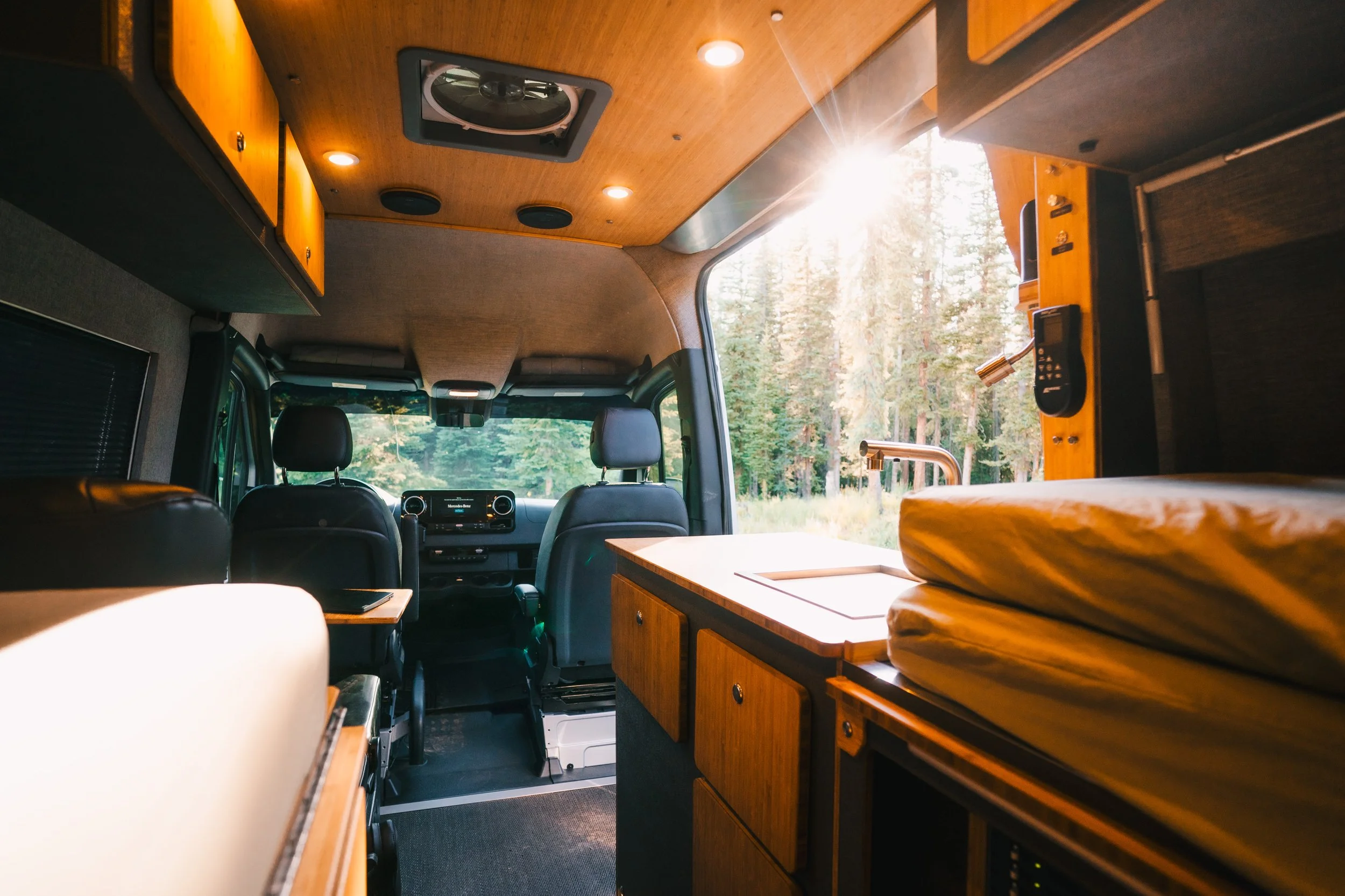 Interior of a camper van with wooden cabinetry, a bed, and seating area, with sunlight streaming through the window onto a forested landscape outside.