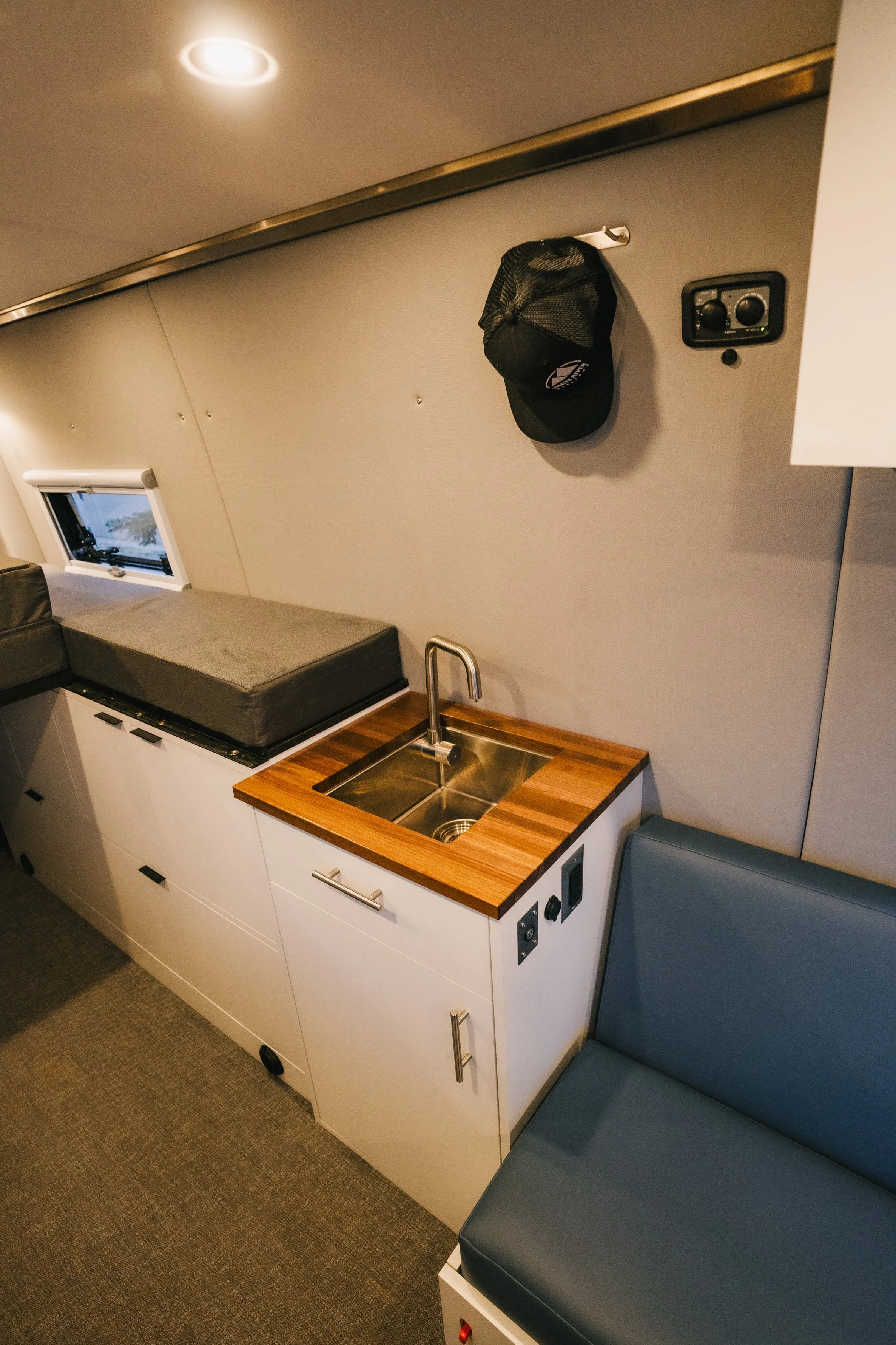 A small kitchen area inside a camper or tiny home, featuring a stainless steel sink with wooden countertop, white cabinetry, a small window, a gray cushioned seat, and a black cap hanging on the wall.