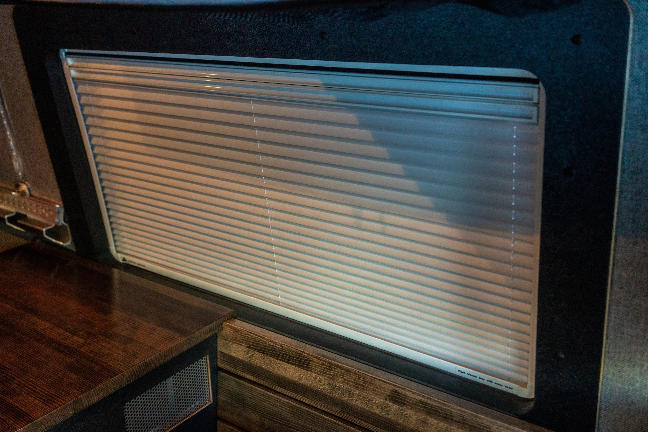 Air vent with closed beige blinds mounted on a black wall next to a wooden table.