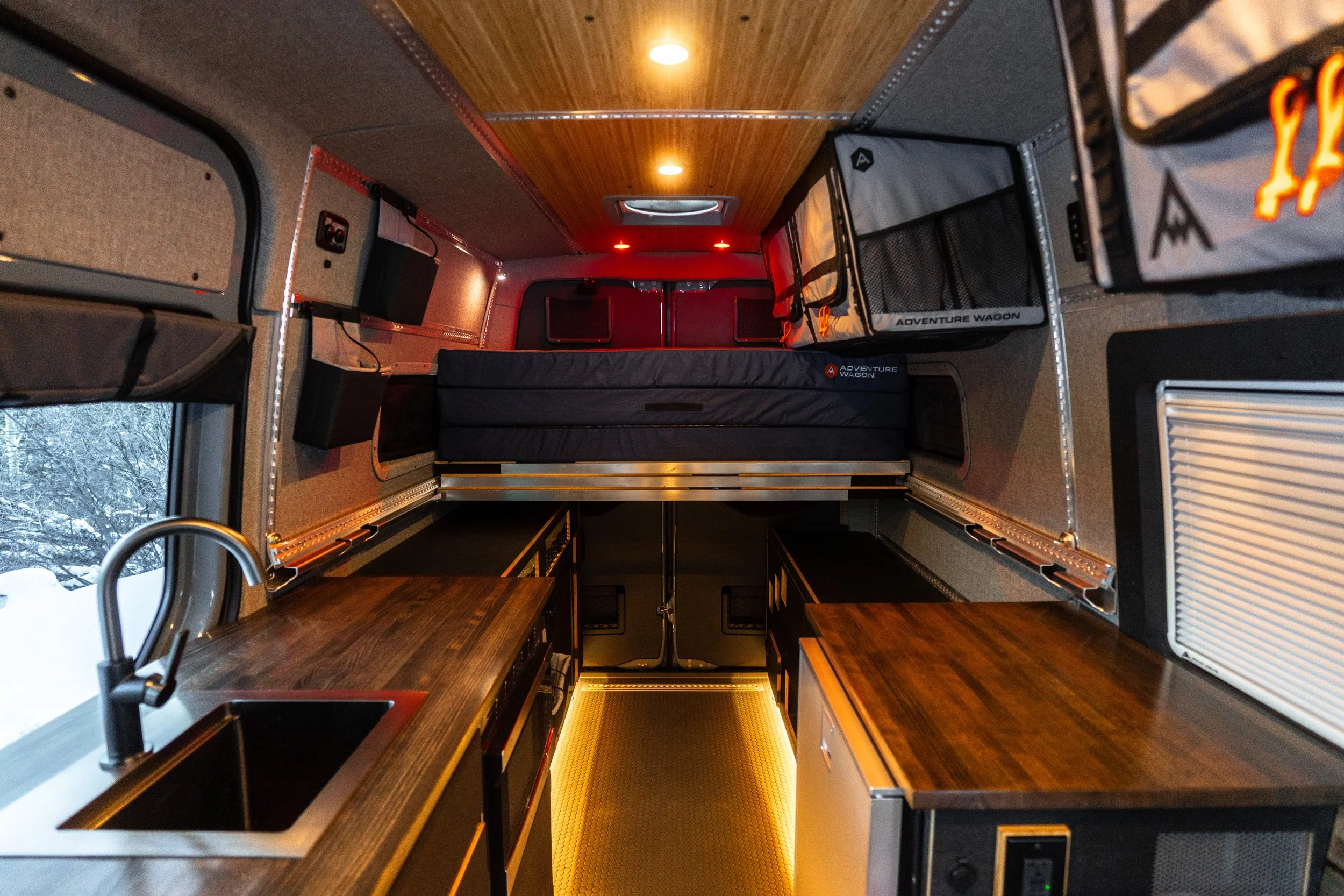 Interior of a camper van with a small kitchen area, wood countertops, a sink, wall-mounted storage, a small fridge, and a sleeping area with a mattress and storage compartments.