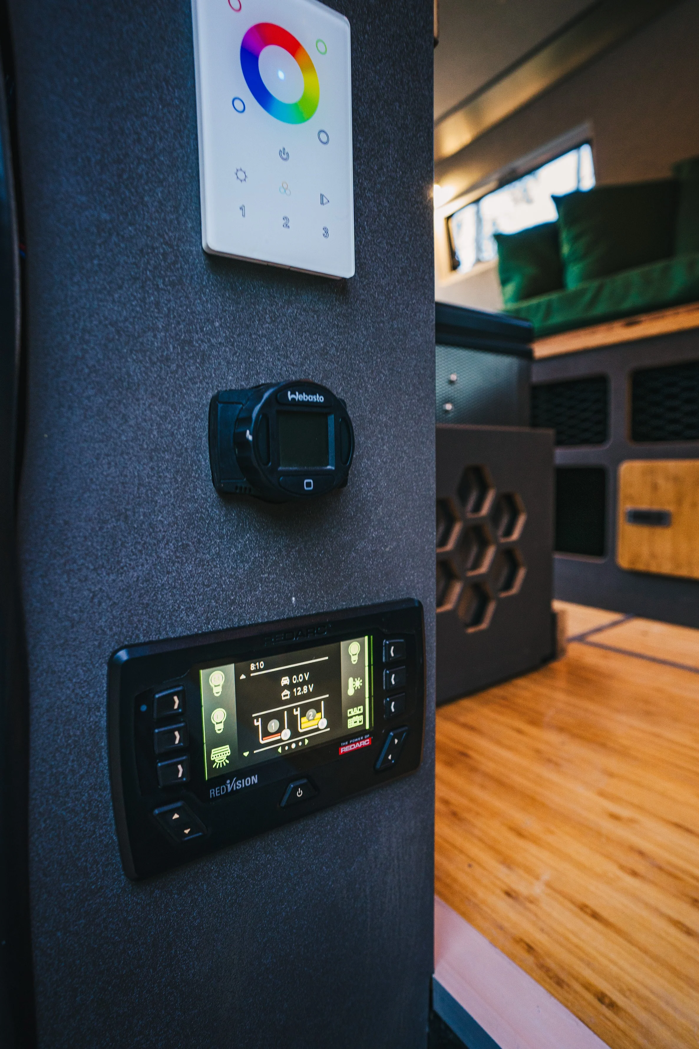 Close-up of control panels on a dark grey wall inside a recreational vehicle, featuring a color-changing LED light control, a digital battery monitor, and a device labeled 'Webasto' that appears to be a thermostat or climate control system.