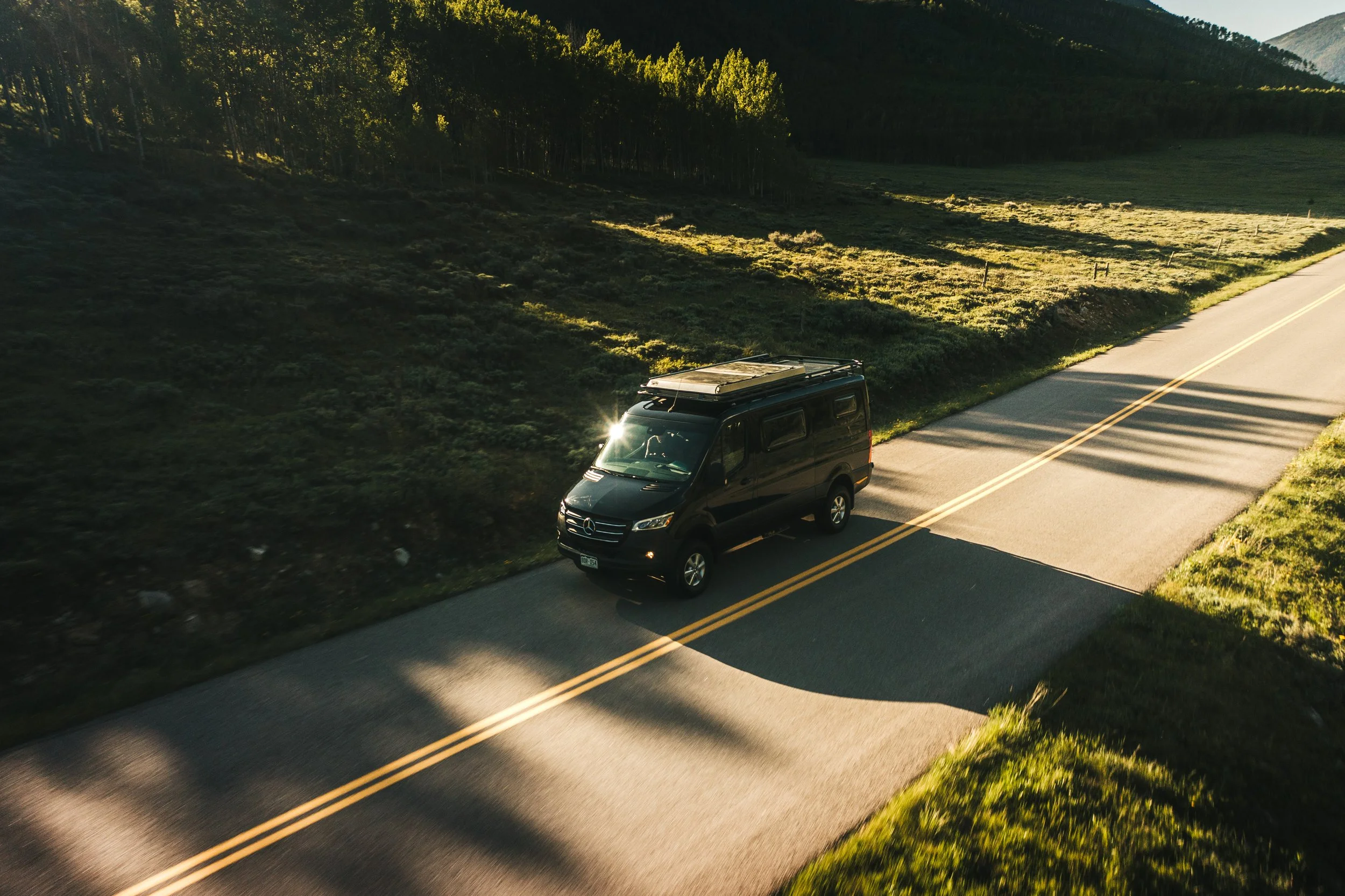 A black Mercedes-Benz van driving on a two-lane rural road surrounded by green grass and trees, with long shadows cast by the setting sun.