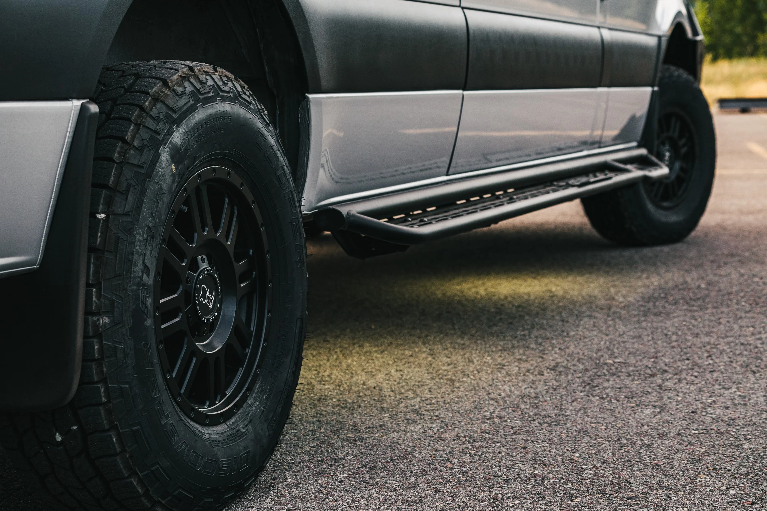 Close-up of the front left side of a black SUV with large off-road tires, black rims, and side steps, parked on a paved road with greenery in the background.