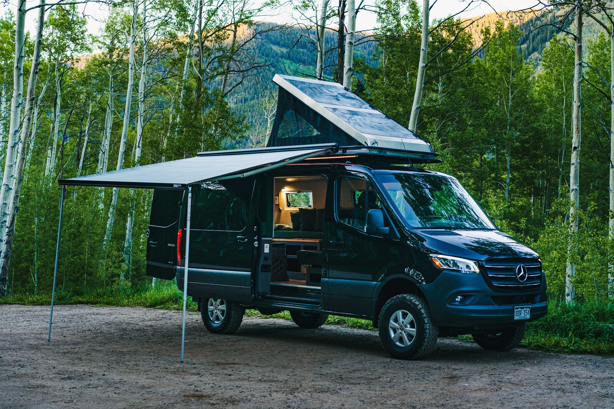 Black camper van with an extended pop-up roof and awning, parked in a forested area with tall trees and mountains in the background.