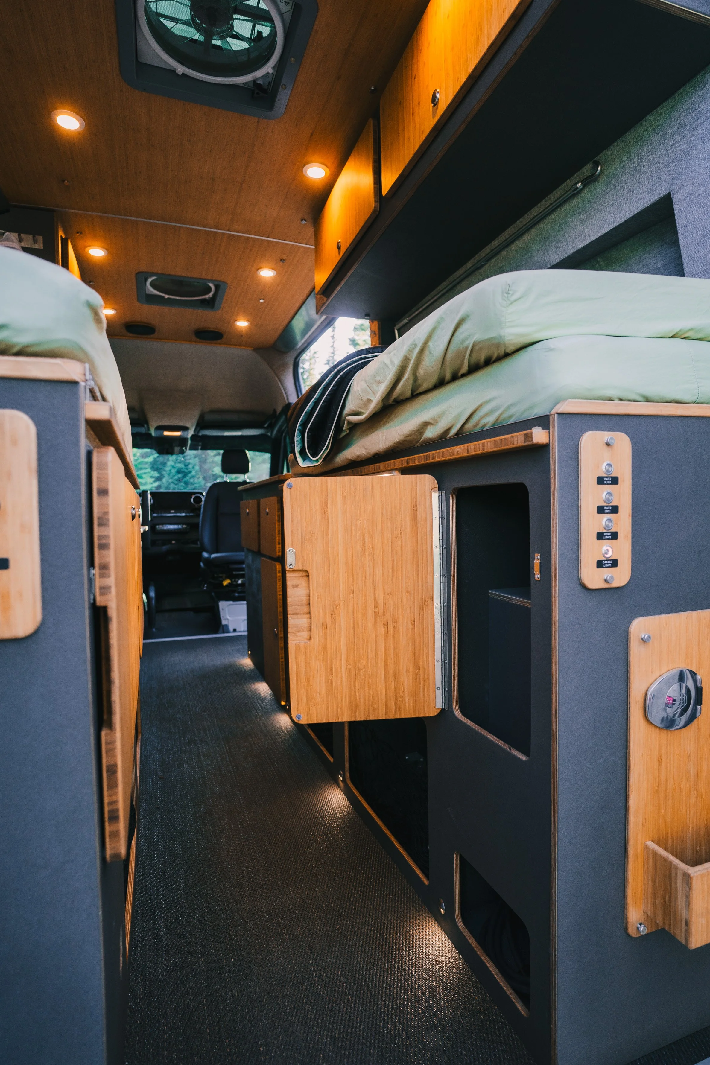 Interior of a camper van with wooden cabinetry, sleeping area with green bedding, and driving seat visible at the front.