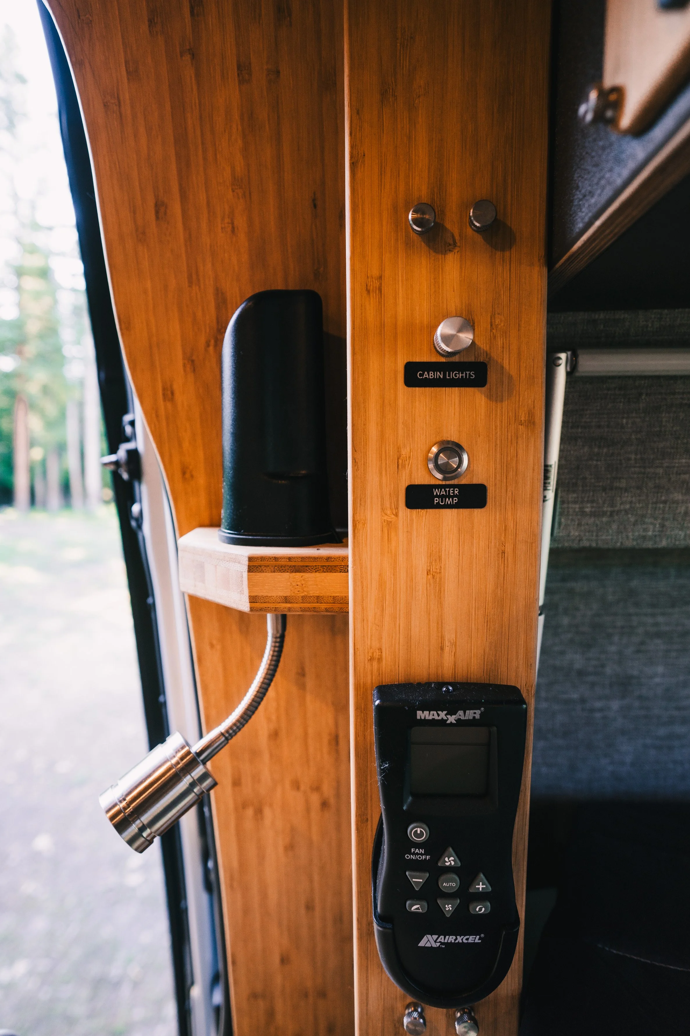 Control panel in a camper with switches labeled 'Cabin Lights' and 'Water Pump', a black speaker, and a remote control for a MaxxAir ceiling fan.