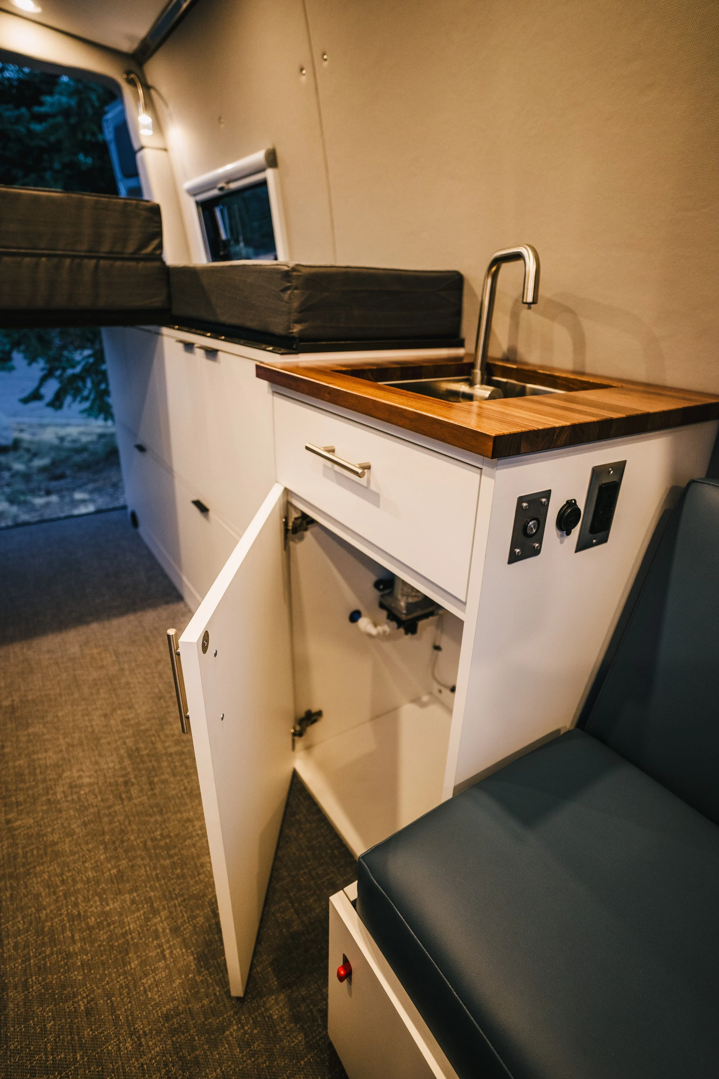 Interior of a camper van showing a small sink with a wooden countertop, storage cabinet with an open door, and a cushioned seating area.