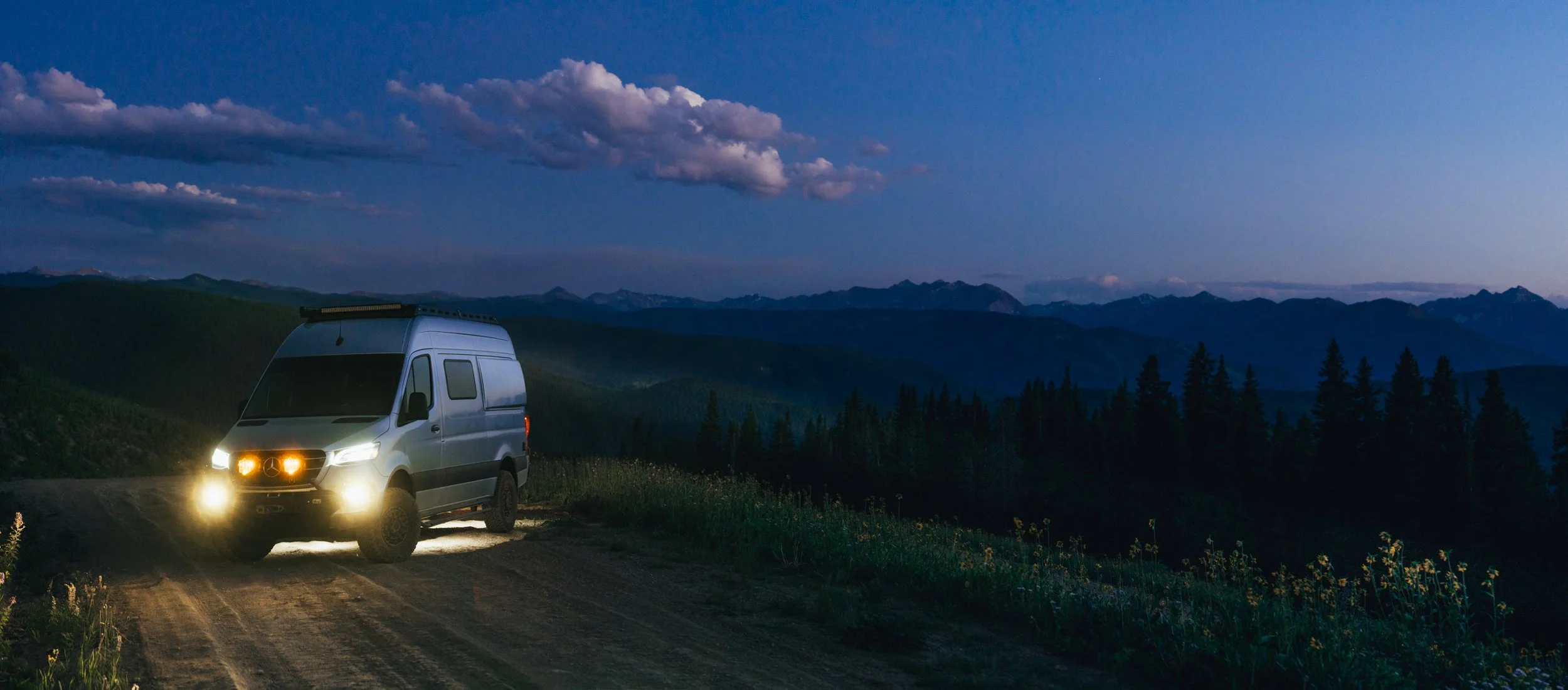 A silver van with bright headlights on a dirt road in a mountainous area at dusk, with a forest and mountains in the background.