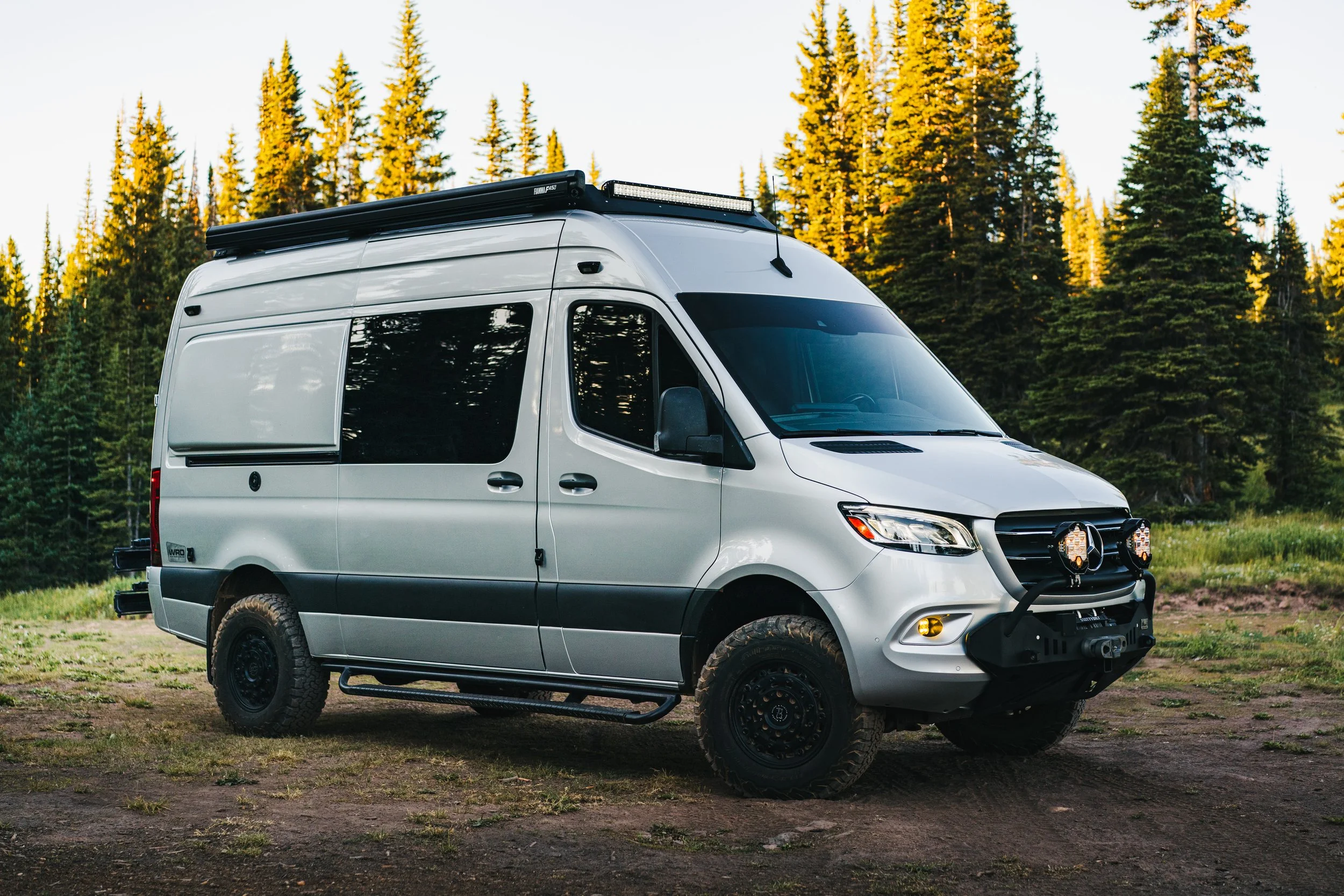 White adventure van with off-road tires and roof-mounted light bar parked on dirt ground in a forested area.