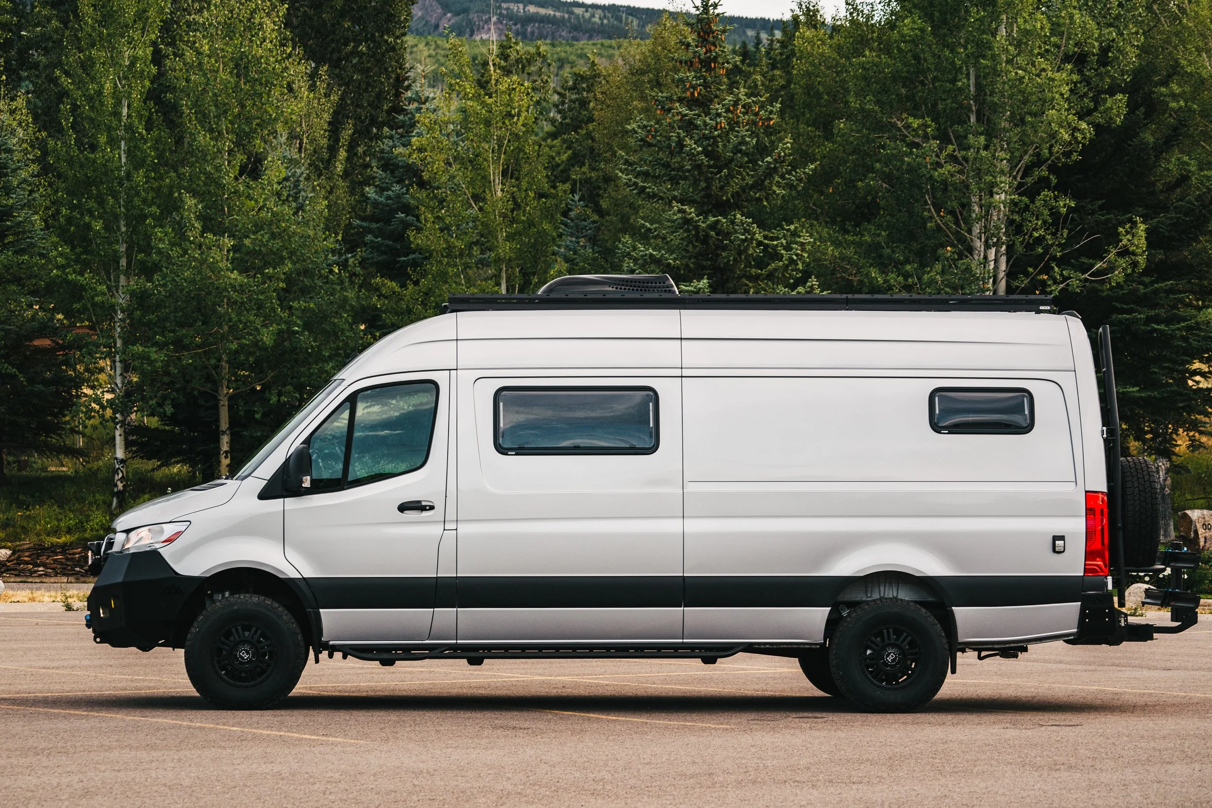 Silver Sprinter camper van parked on a paved surface with a backdrop of green trees and a mountainous landscape.