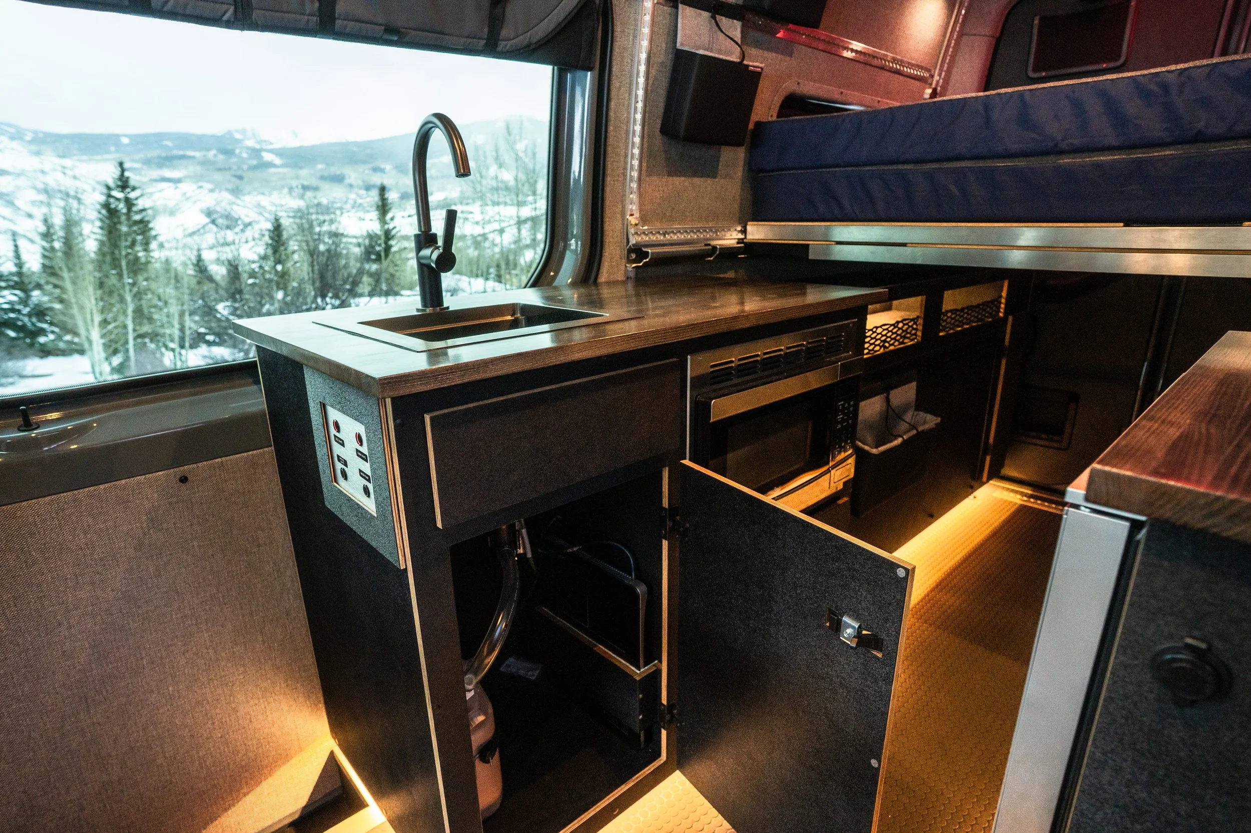 Interior of a compact camper van with a small kitchen area featuring a sink, microwave, and storage cabinets, with a view of snowy mountains and trees through the window.