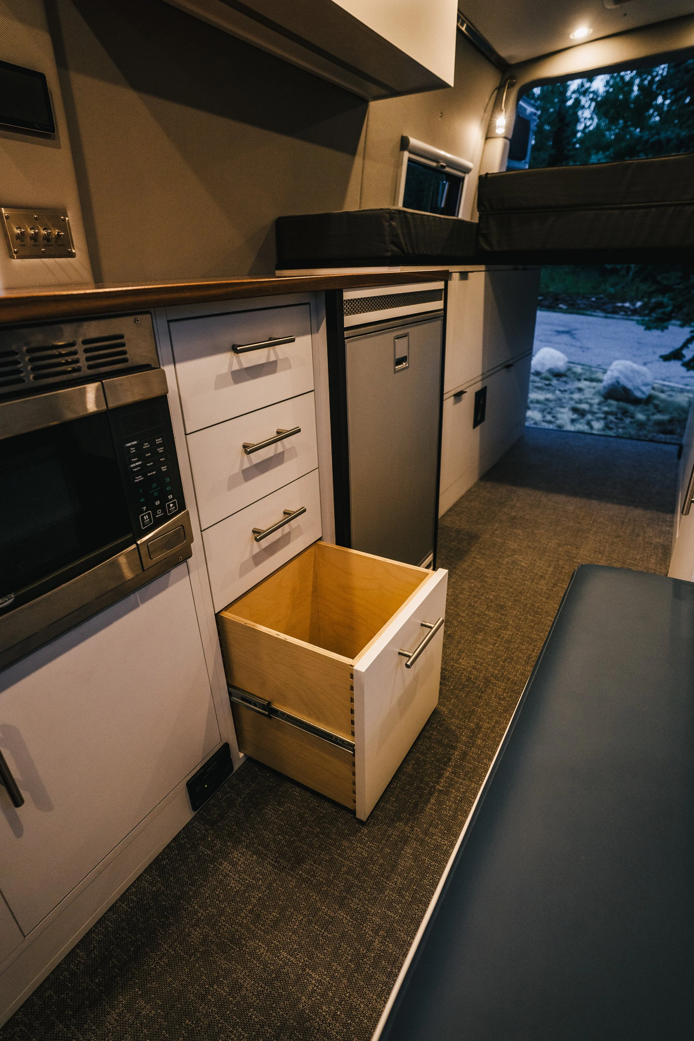 Interior of a camper van kitchen with drawers, a microwave, a mini fridge, an open drawer, and a window showing an outdoor scene with rocks.