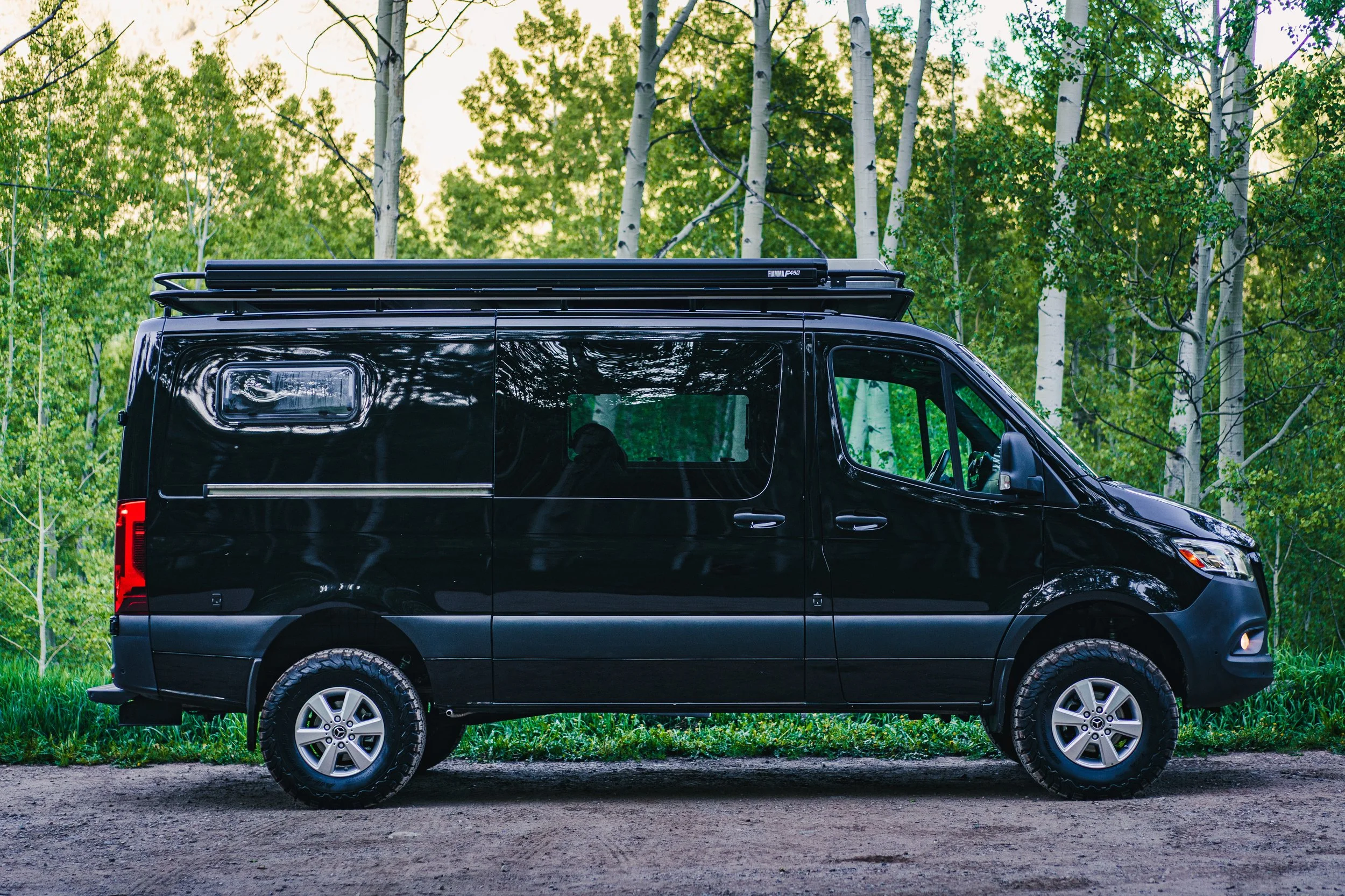 A black van parked on a dirt road in a forest with green trees and birch trees in the background.