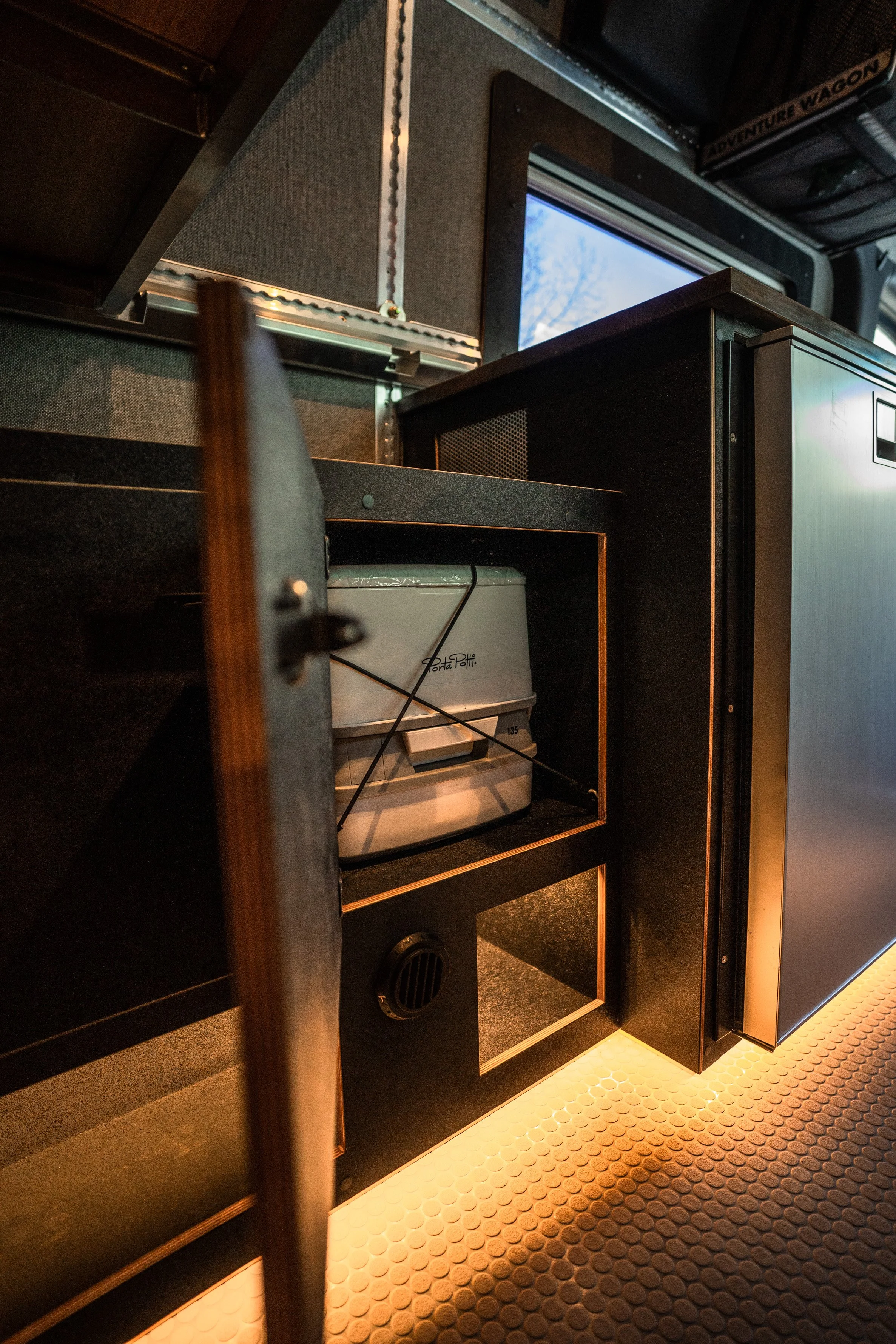 Open cabinet door with a portable oven or toaster oven inside on a countertop. A window is visible in the background with a tree outside. The setting appears to be part of a vehicle interior, possibly an RV or camper, with textured flooring and shelv