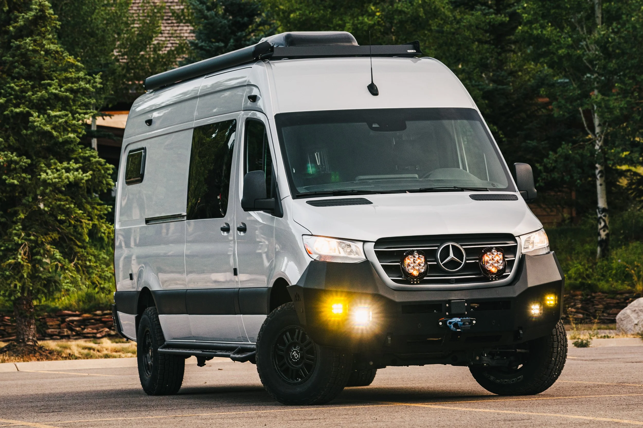 Silver Sprinter camper van with black front bumper and round auxiliary lights on the grille, parked on a paved road with green trees and bushes in the background.
