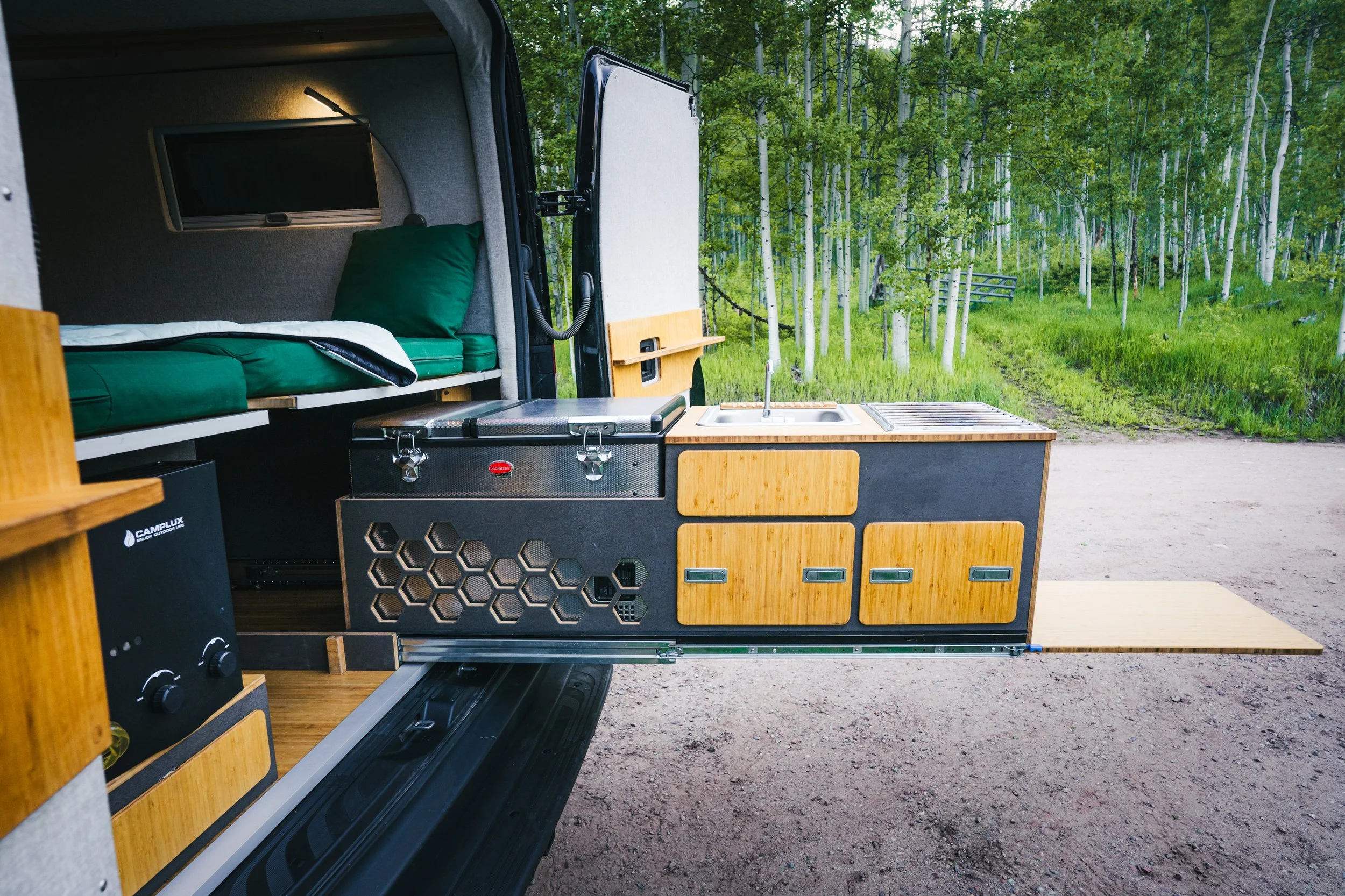 Interior of a camper van with a kitchenette and a sleeping area, set in a forested outdoor location.