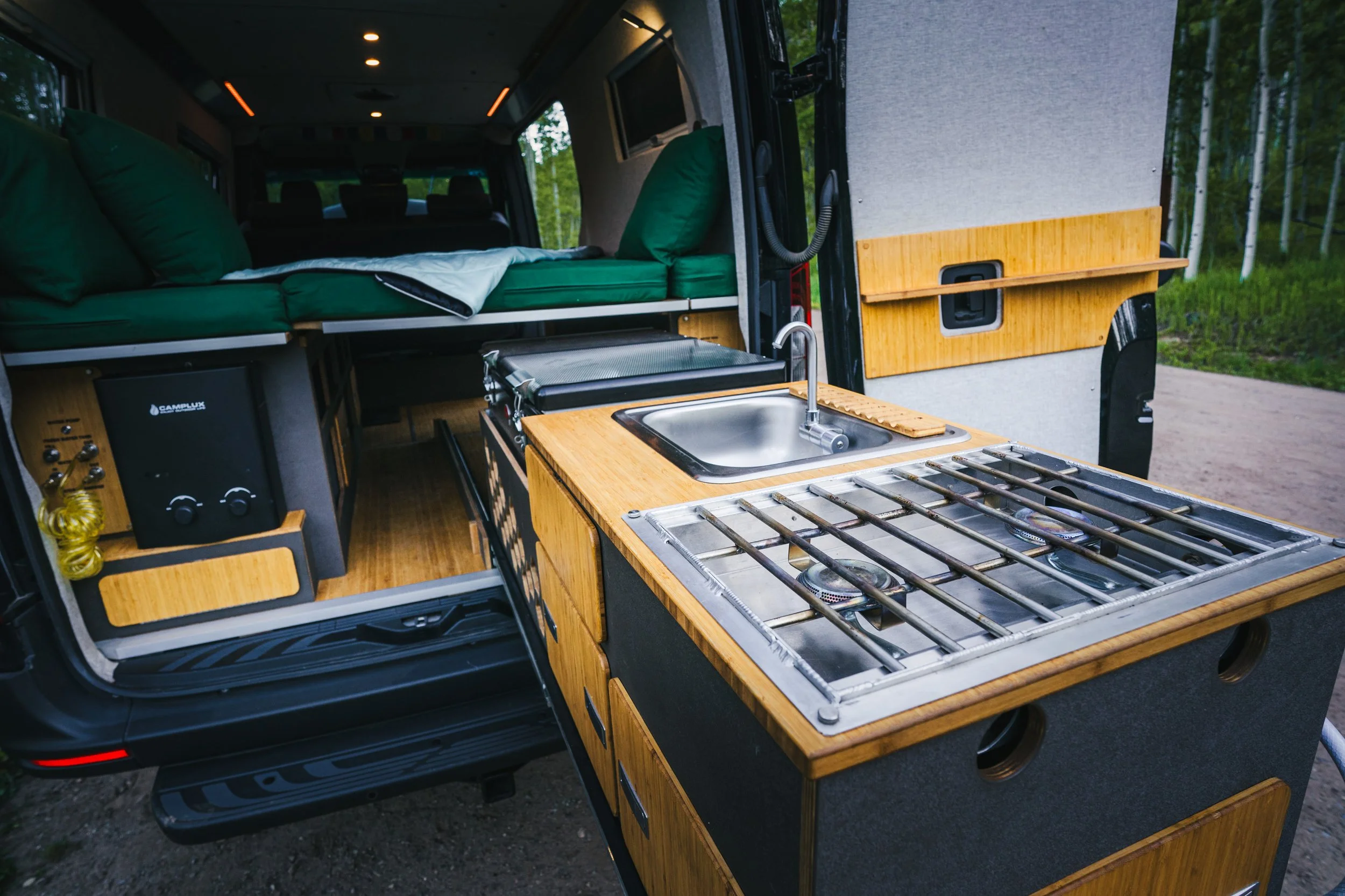 Inside a camper van with a wooden kitchen countertop featuring a sink and a gas stove, green cushions, a bed with green bedding, and a view of trees outside.