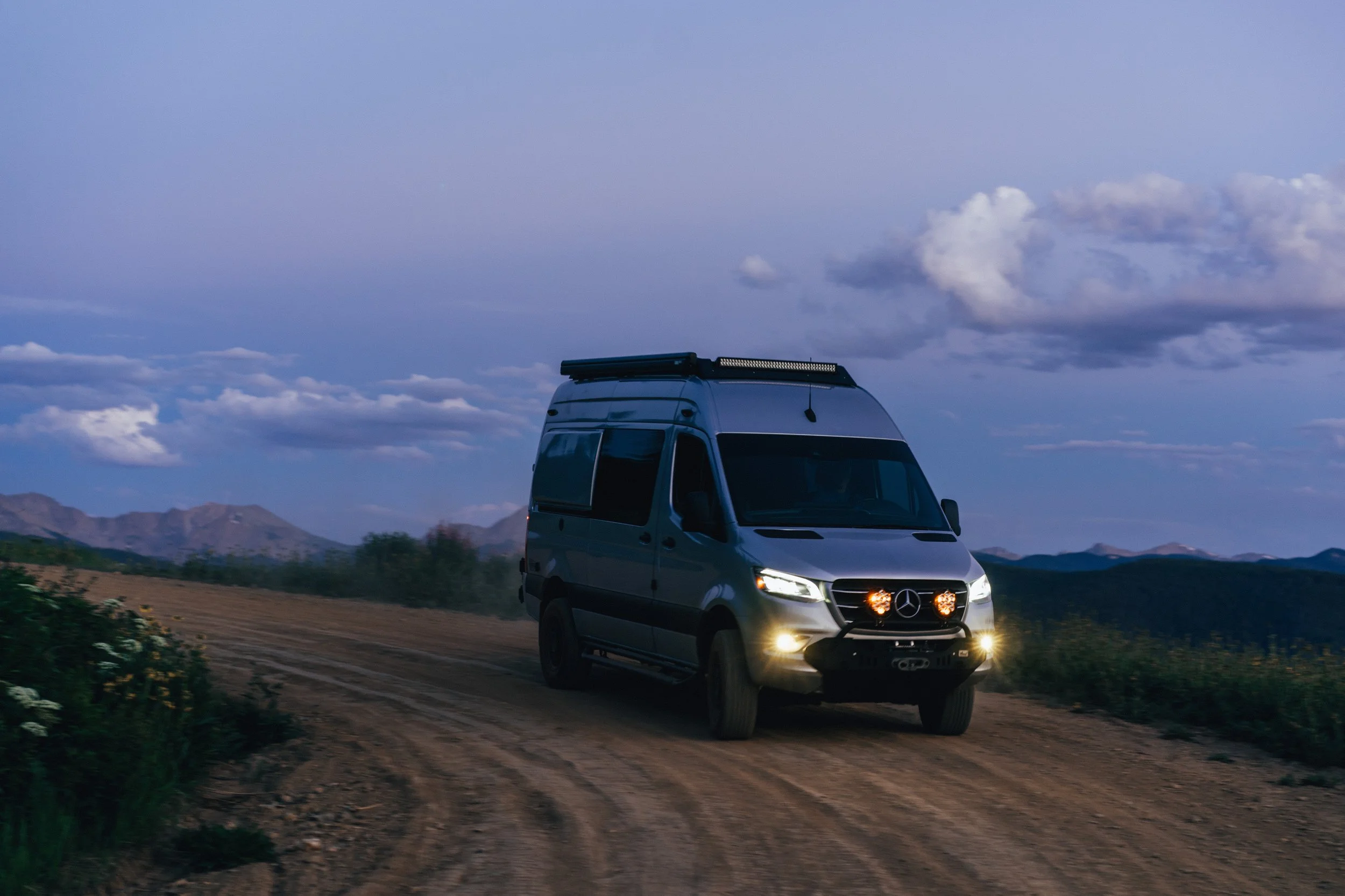 A silver camper van driving on a dirt road through a rural landscape at dusk with mountains and a cloudy sky in the background.