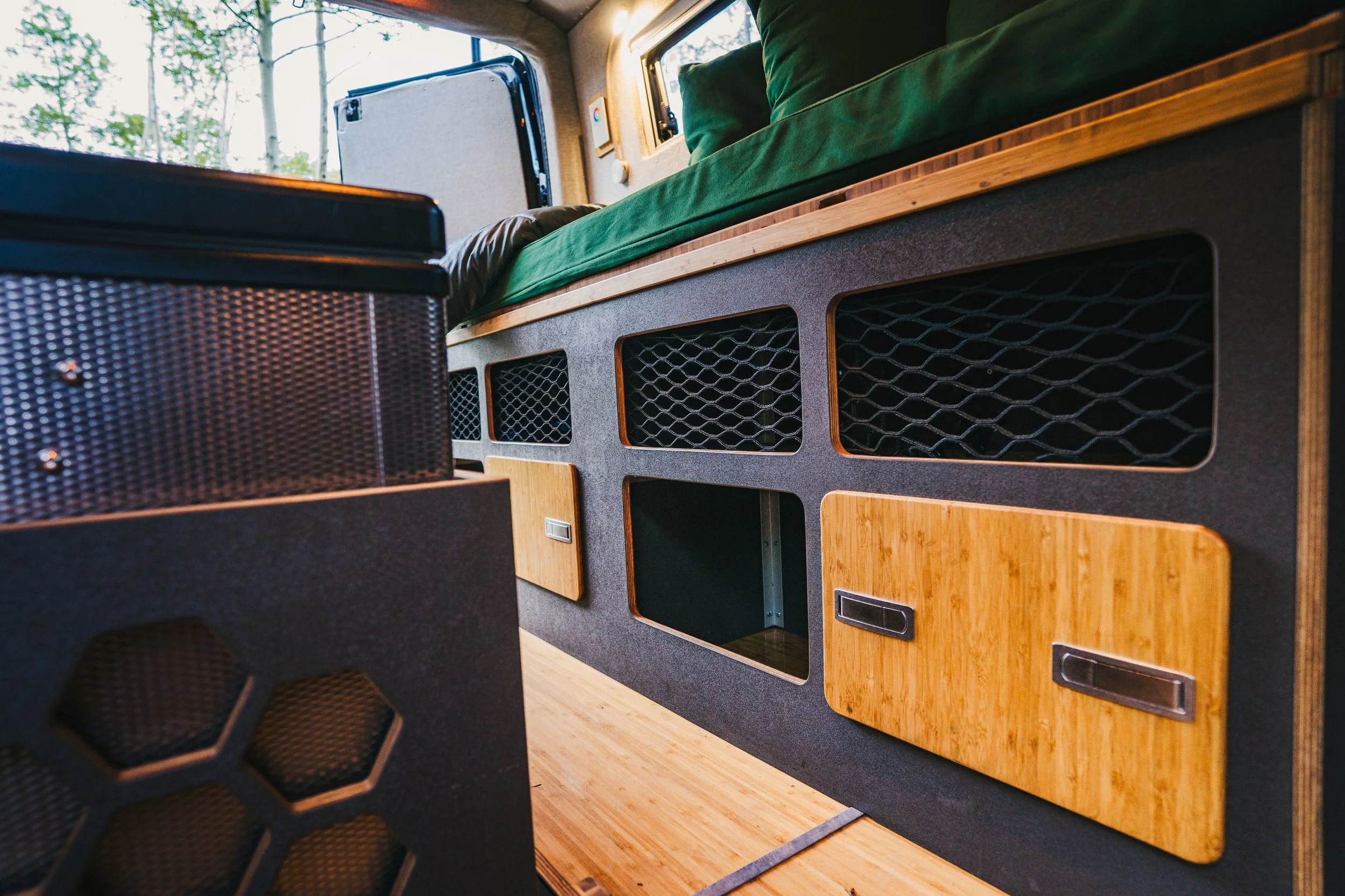 Interior of a camper van with built-in storage cabinets, a bed with green bedding, and a window with trees outside.