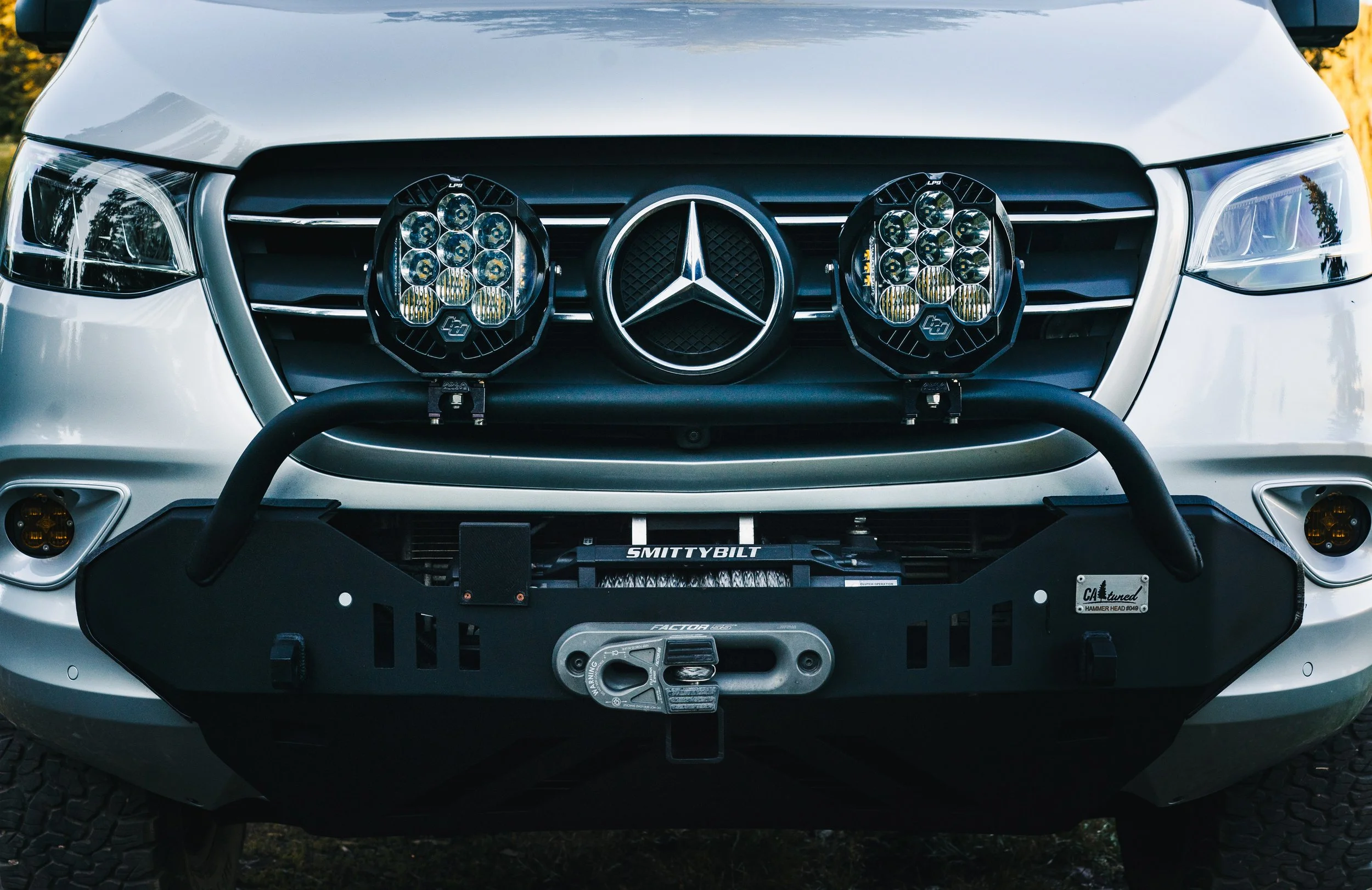 Front view of a silver Mercedes-Benz vehicle with off-road accessories, including a bumper with a mounted winch, two large round floodlights, and a grille featuring the Mercedes-Benz emblem.