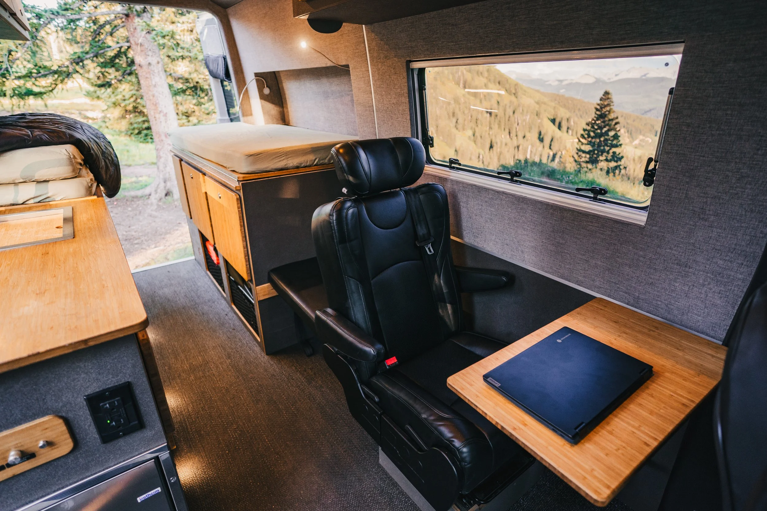 Interior of a camper van with a black leather seat, a wooden table with a closed laptop, and a sleeping area with a bed near the window showing mountain and forest scenery.