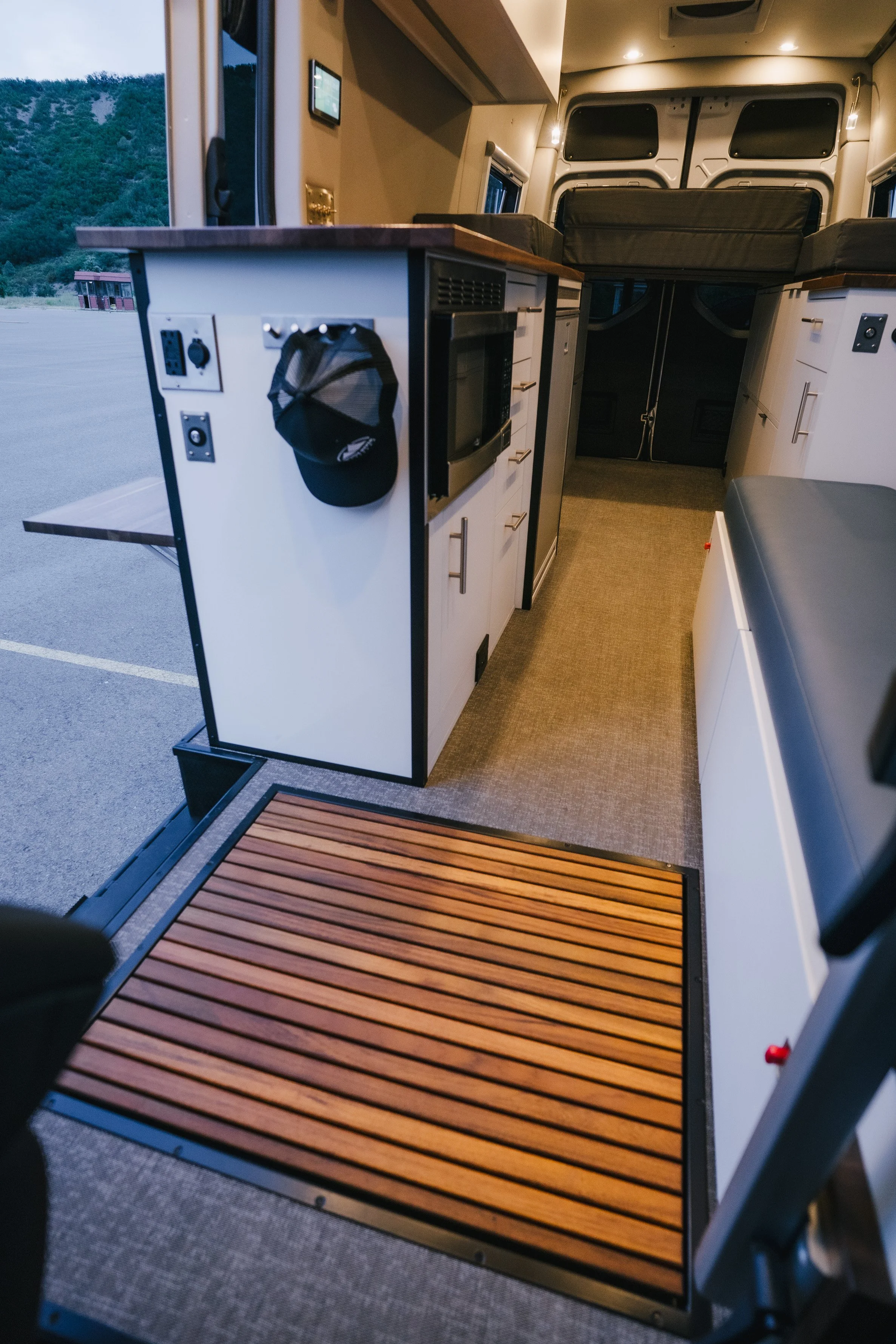 Interior of a camper van with small kitchen area, including a microwave, countertop, under-counter refrigerator, and a bed in the back, with a mountain view outside the open side door.