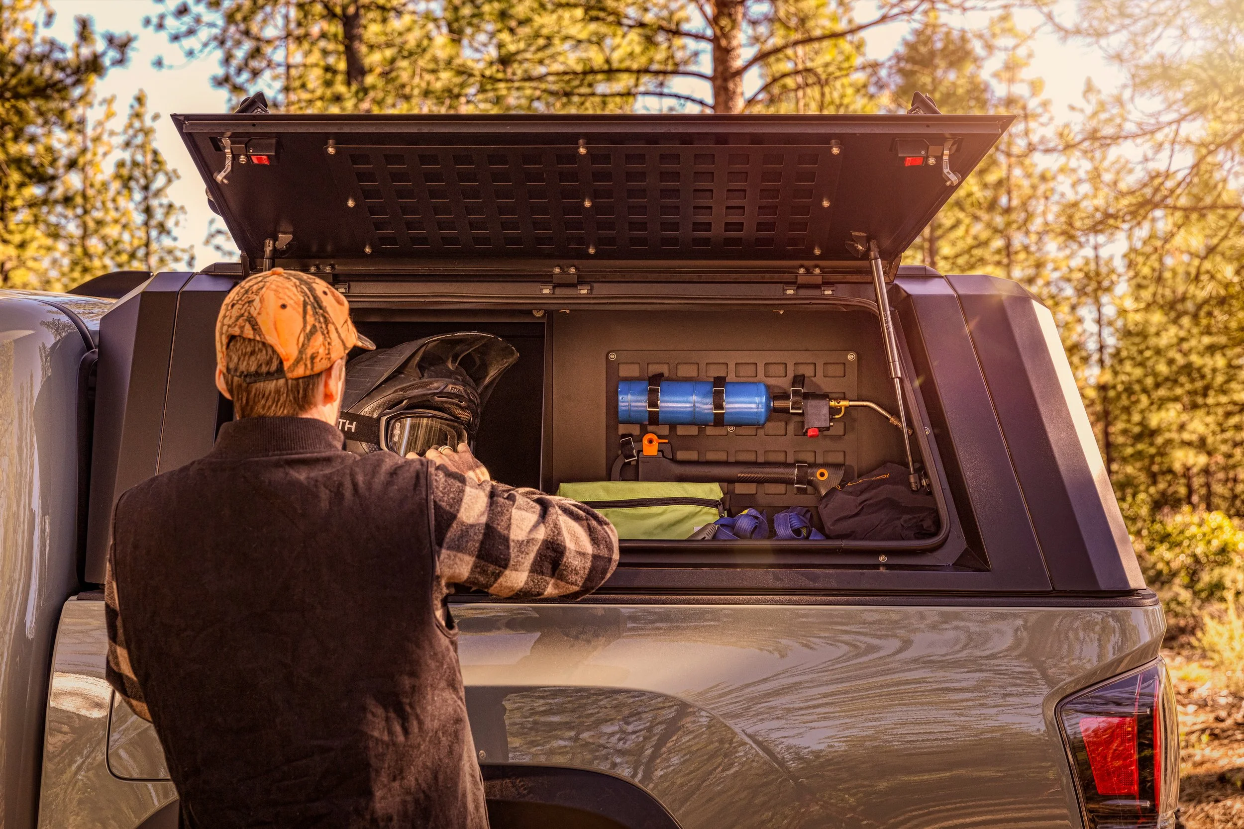 A person with a camouflage cap and checkered shirt putting on a helmet near the open bed of a pickup truck, which is equipped with gear and tools for outdoor activities, in a wooded area during sunset.