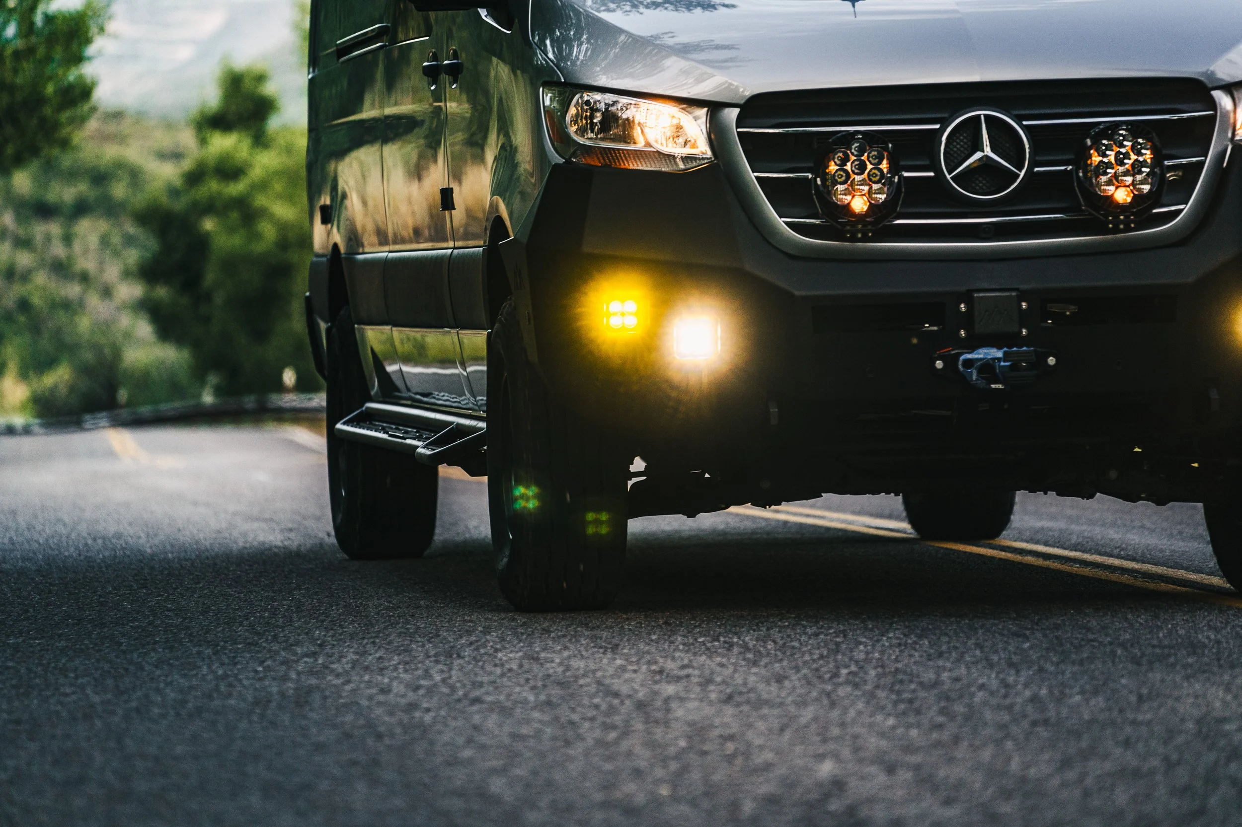 Close-up of the front of a silver Mercedes-Benz SUV driving on a two-lane road with greenery in the background, with its headlights and additional LED lights turned on.