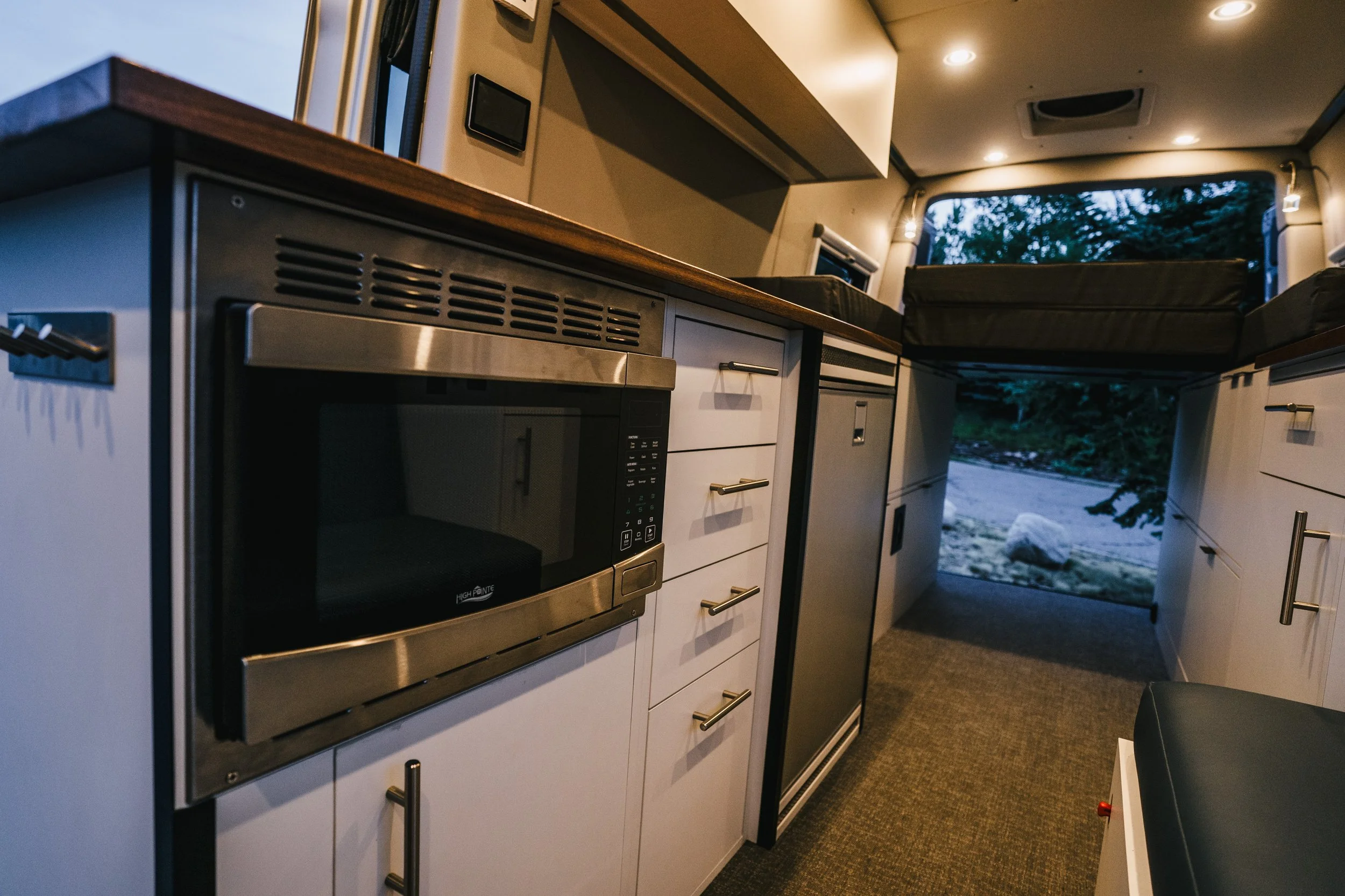 Interior of a compact RV kitchen with white cabinets, a microwave, and a view to the outside through an open rear door showing a natural landscape.