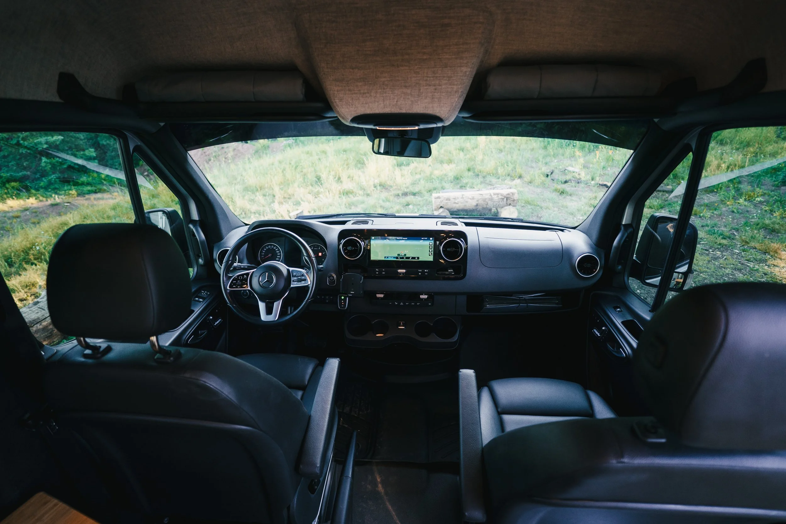 Interior view of a Mercedes-Benz camper van with black leather seats, A navigation screen, and a dashboard, parked in a natural setting with greenery visible through the windshield.