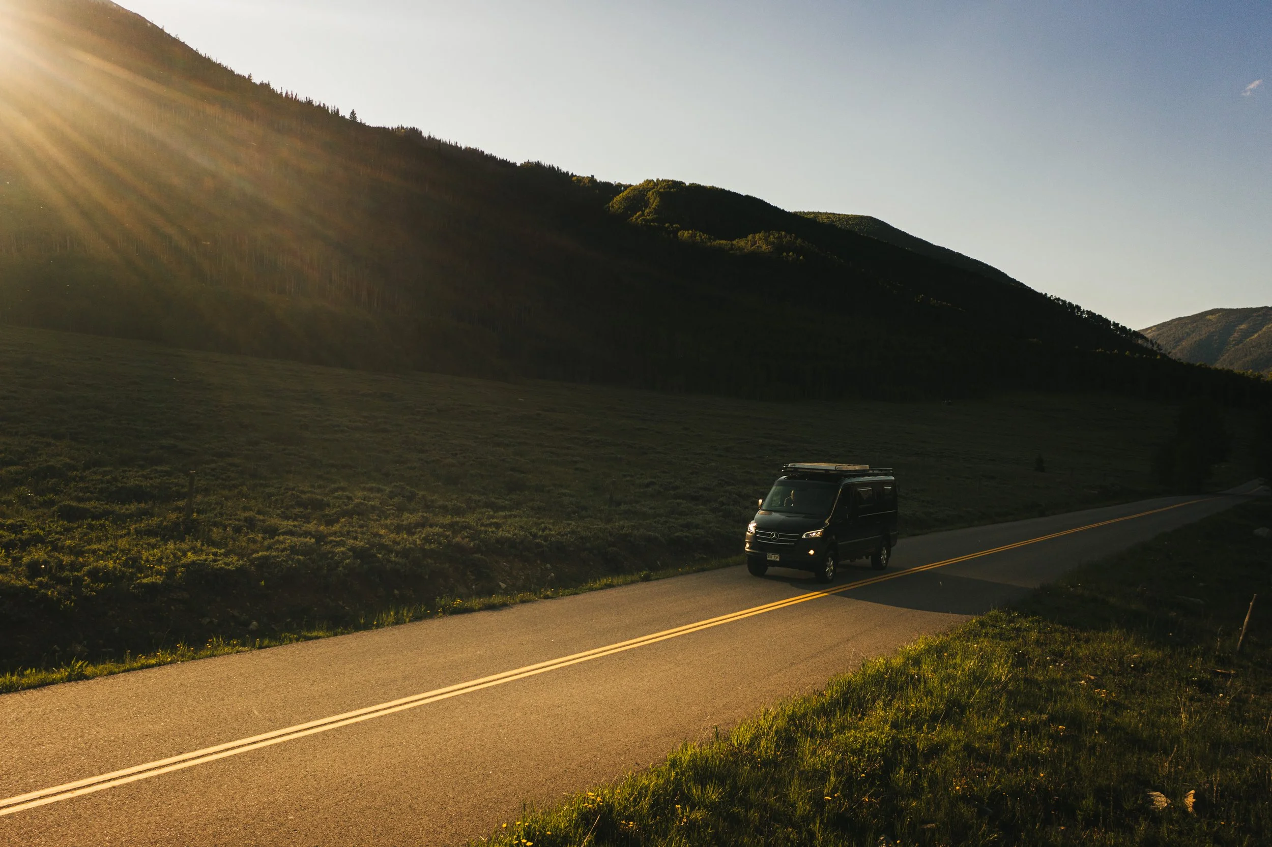 A black van driving on a rural road surrounded by green fields and mountains at sunset.