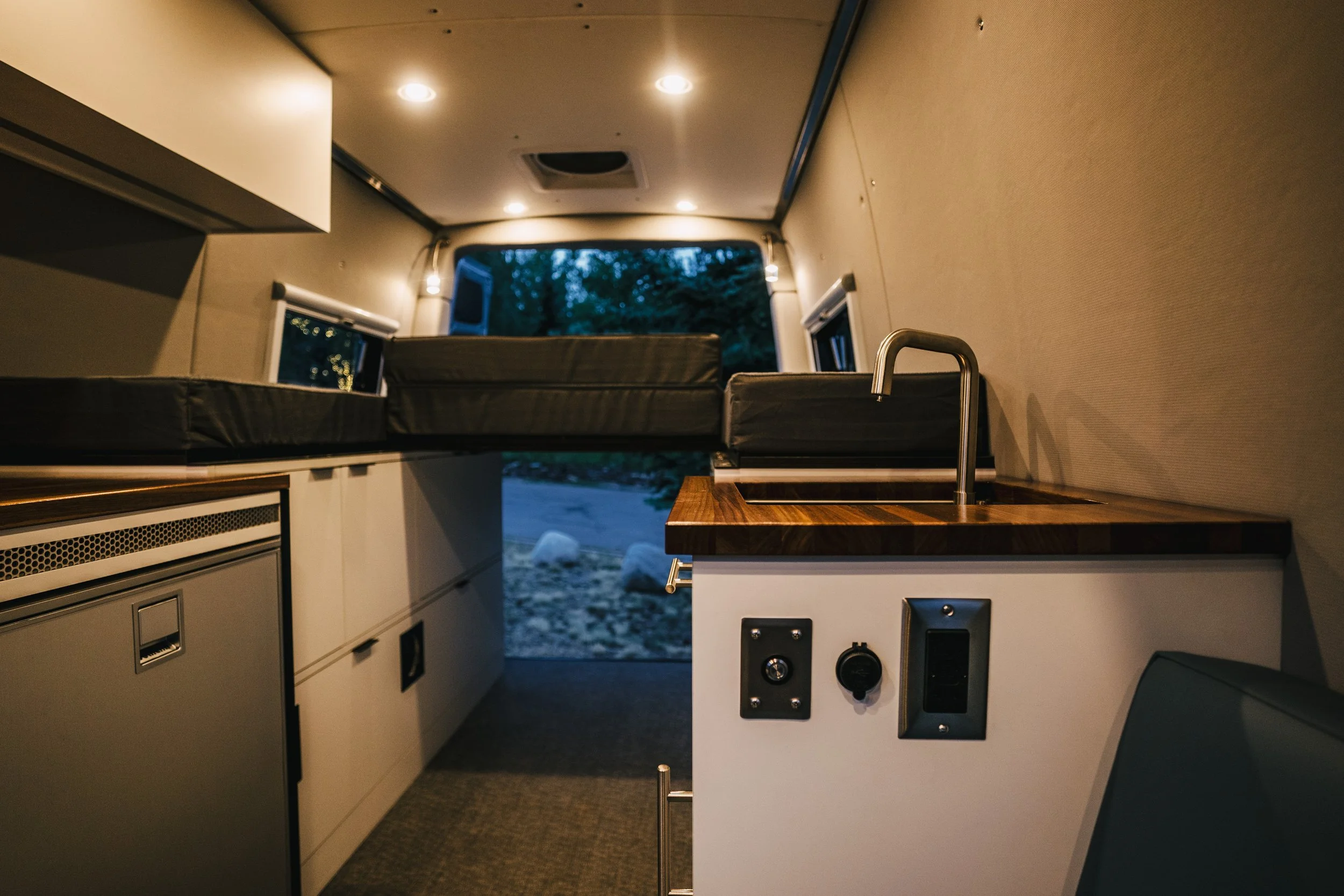 Interior of a converted van or tiny home with a built-in bed, small kitchenette, and window views of trees outside, dimly lit with ceiling lights.