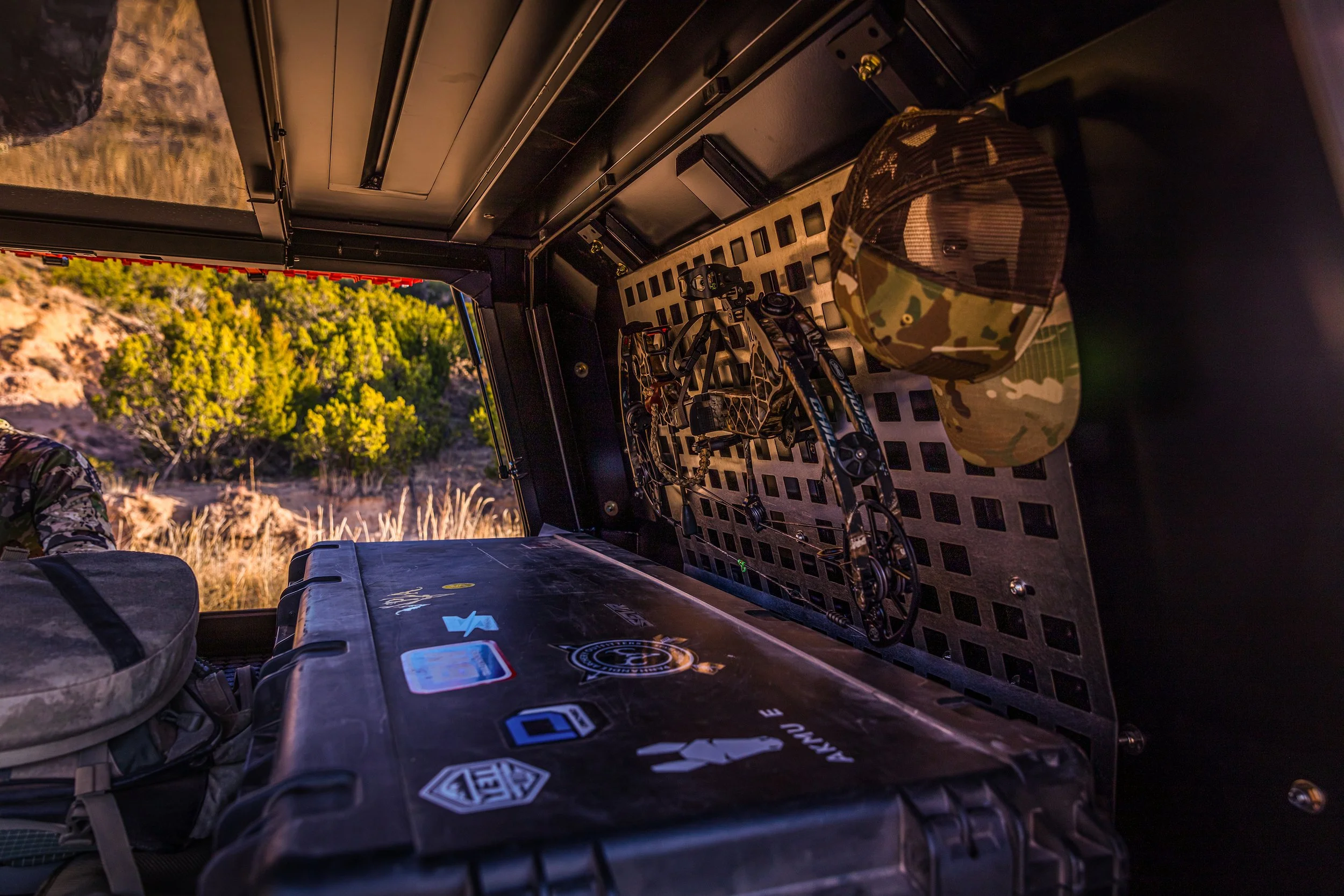 Interior of a vehicle, showing a rifle, camouflage hat, and gear storage area, with a scenic desert landscape outside the window.