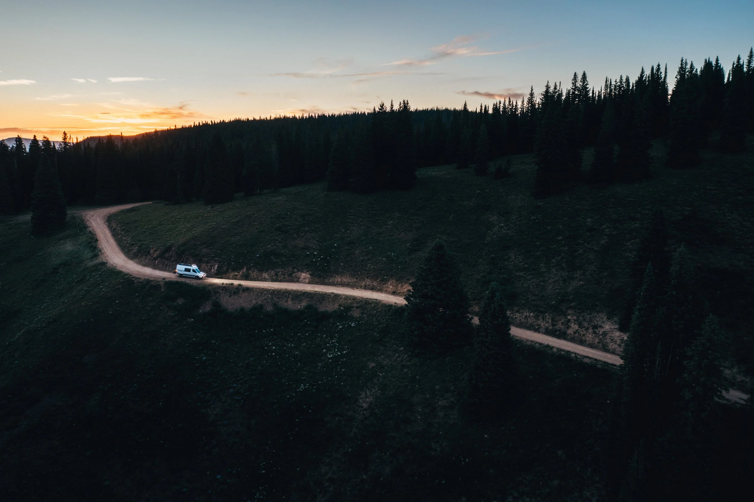 A white van traveling on a winding dirt road through a green hillside with a dense forest of pine trees under a sunset sky.