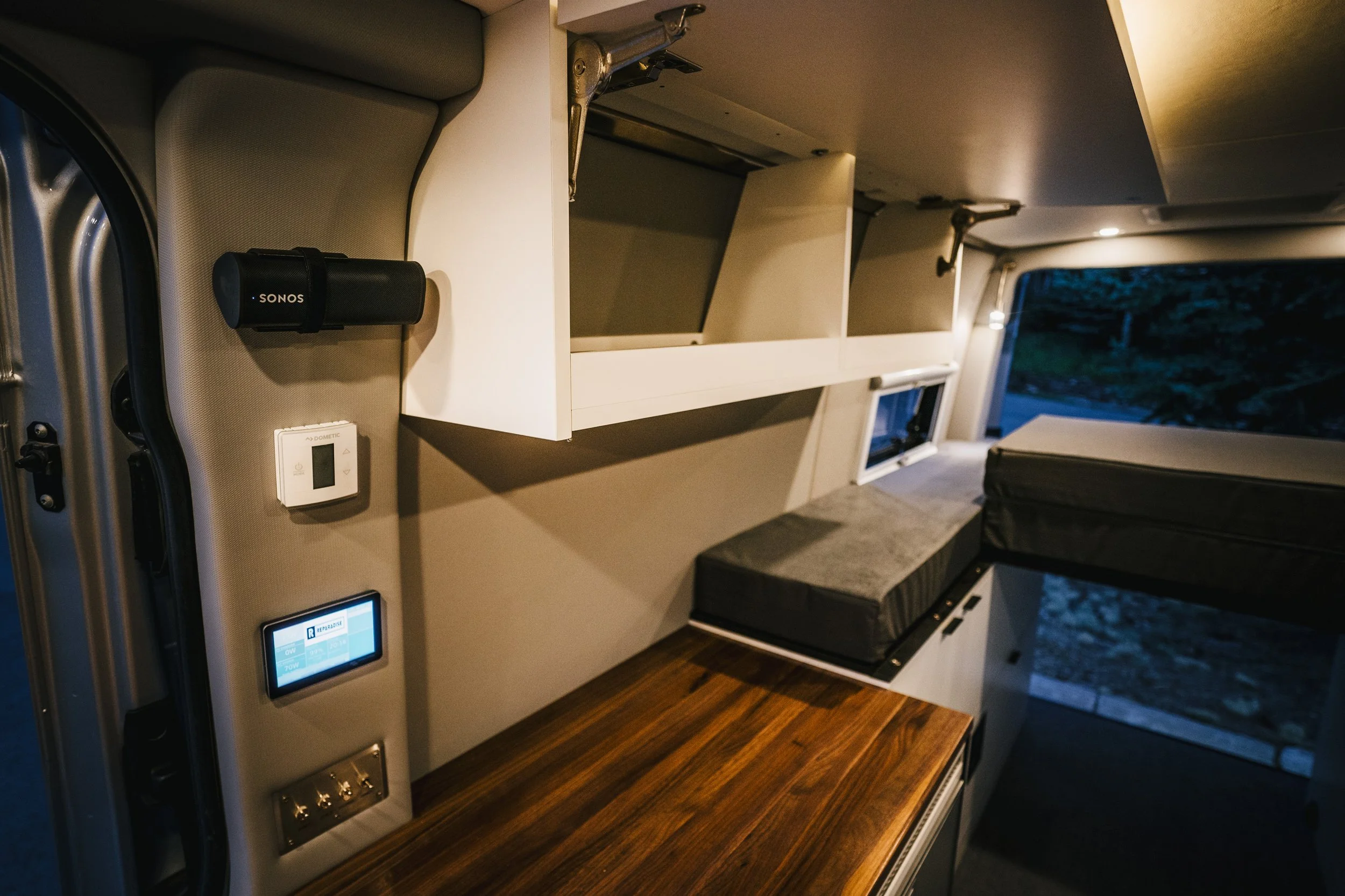 Interior of a camper van with open storage cabinets, a cushioned bench, a moon roof, and electronic control panels.