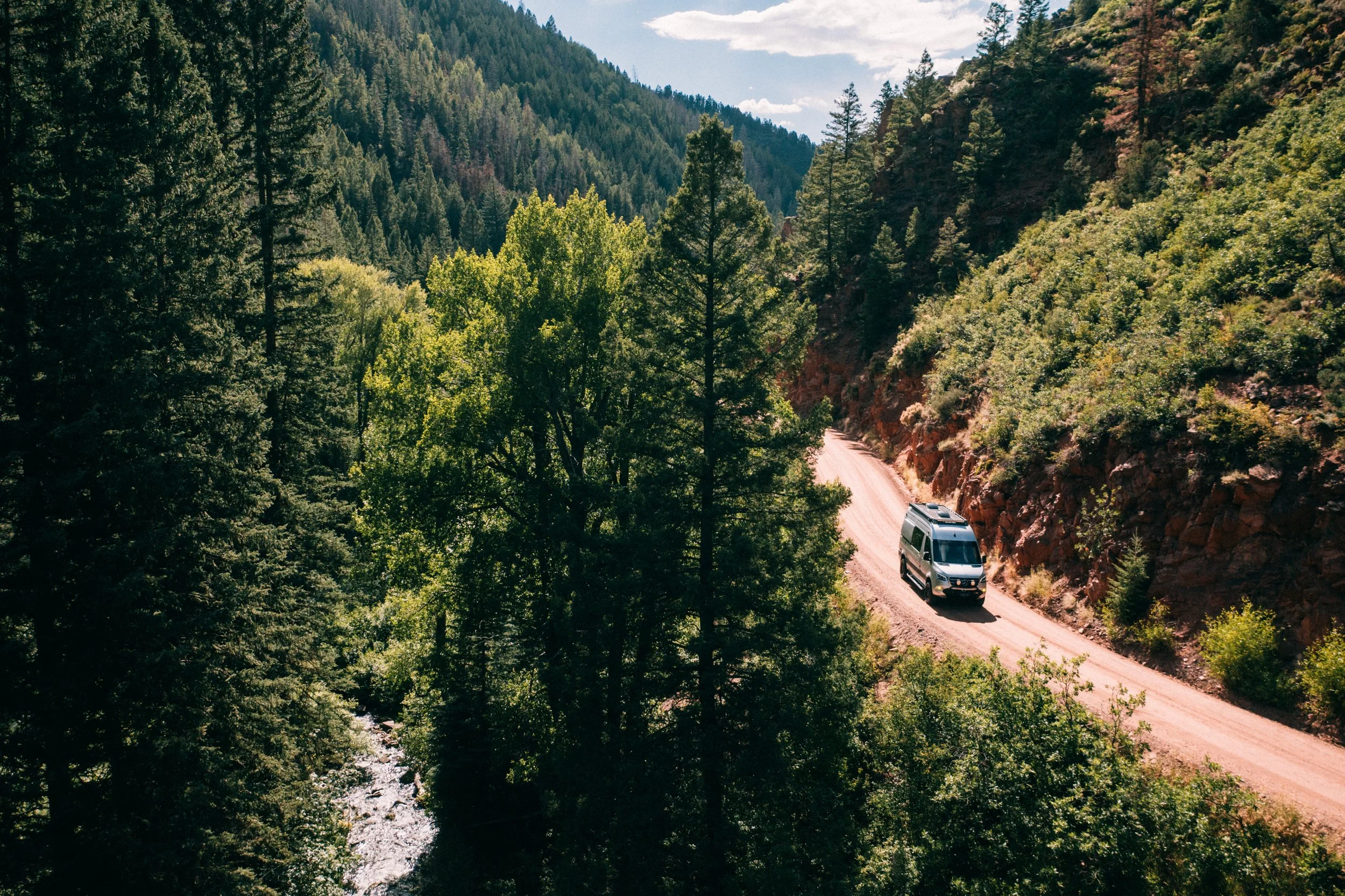 A white van driving on a winding dirt road through a forested mountainous area with tall trees and hills in the background.
