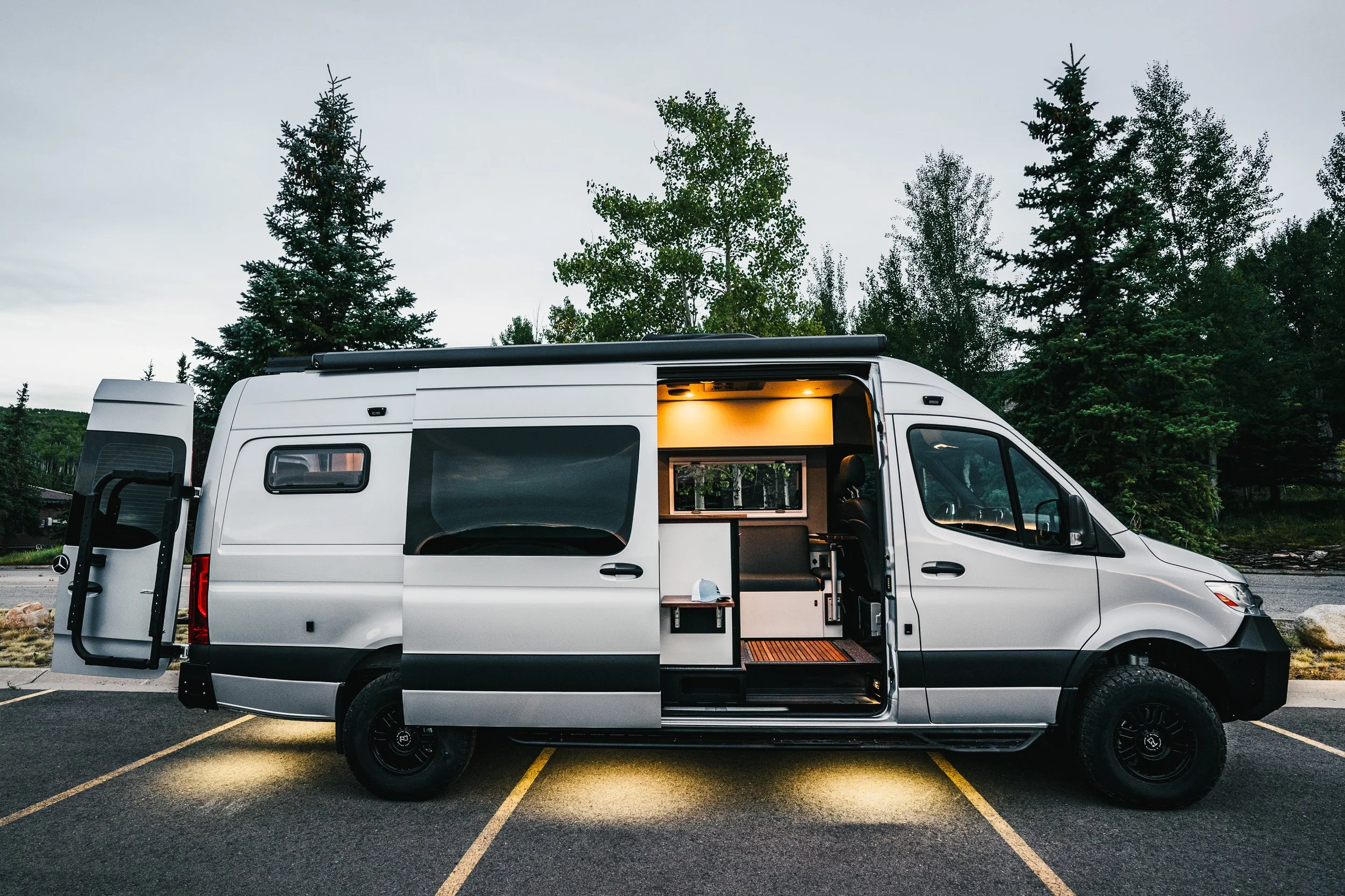 Silver camper van parked in a lot with trees in the background, interior lights on, with open side door showing a small living space.