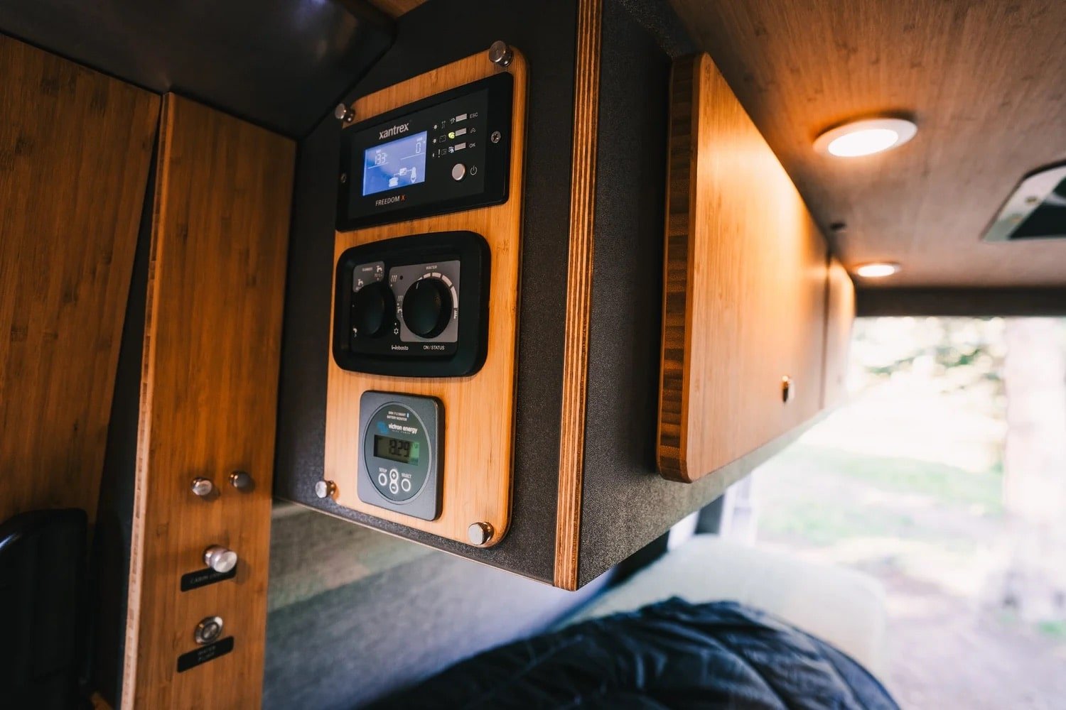 Interior of a camper van showing control panels, including a digital thermostat, a fan control switch, and a digital voltage meter, mounted on wood-paneled walls near a seating area.
