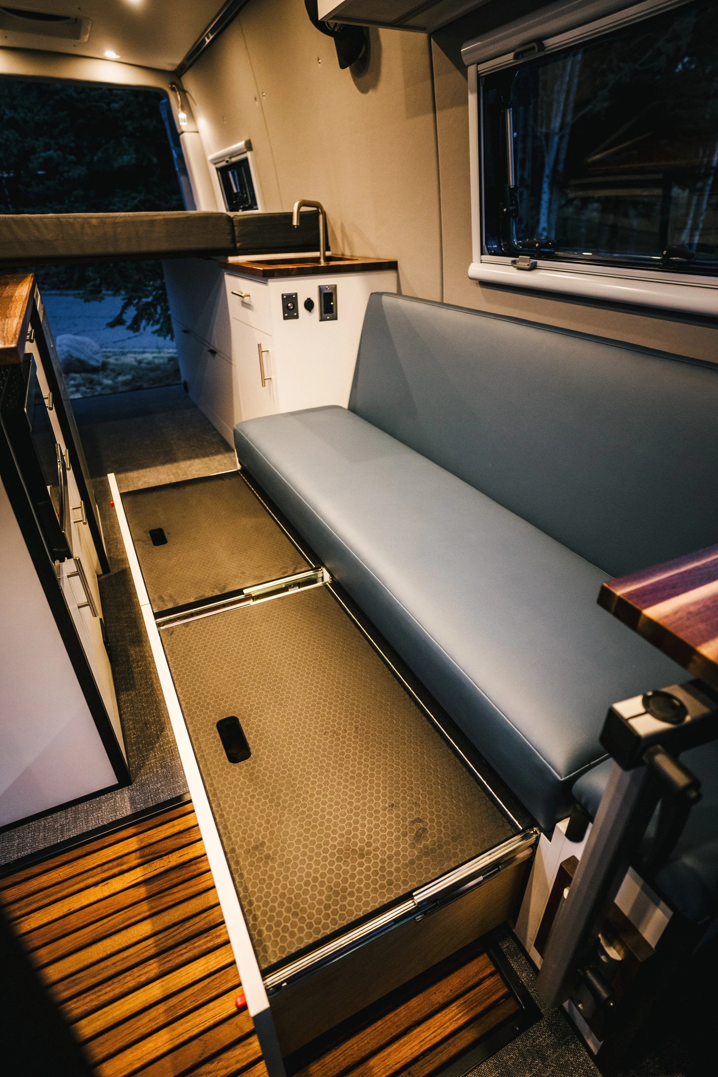 Interior of a camper van with a gray bench seat, a small kitchenette with a sink, and a window showing trees outside.