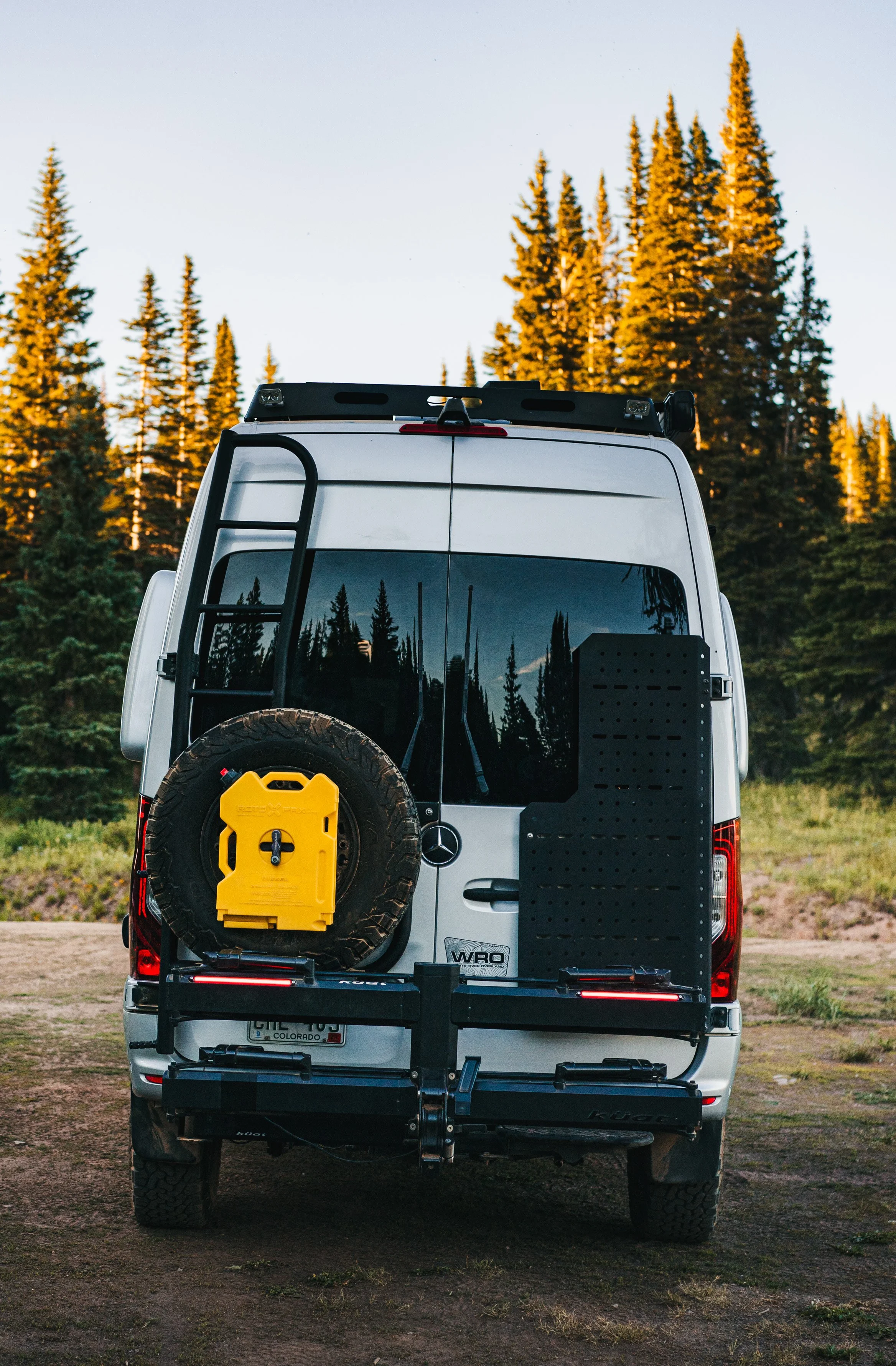 Rear view of a white Mercedes-Benz van with off-road gear and a spare tire mounted on the back, parked in a forested area during sunset.