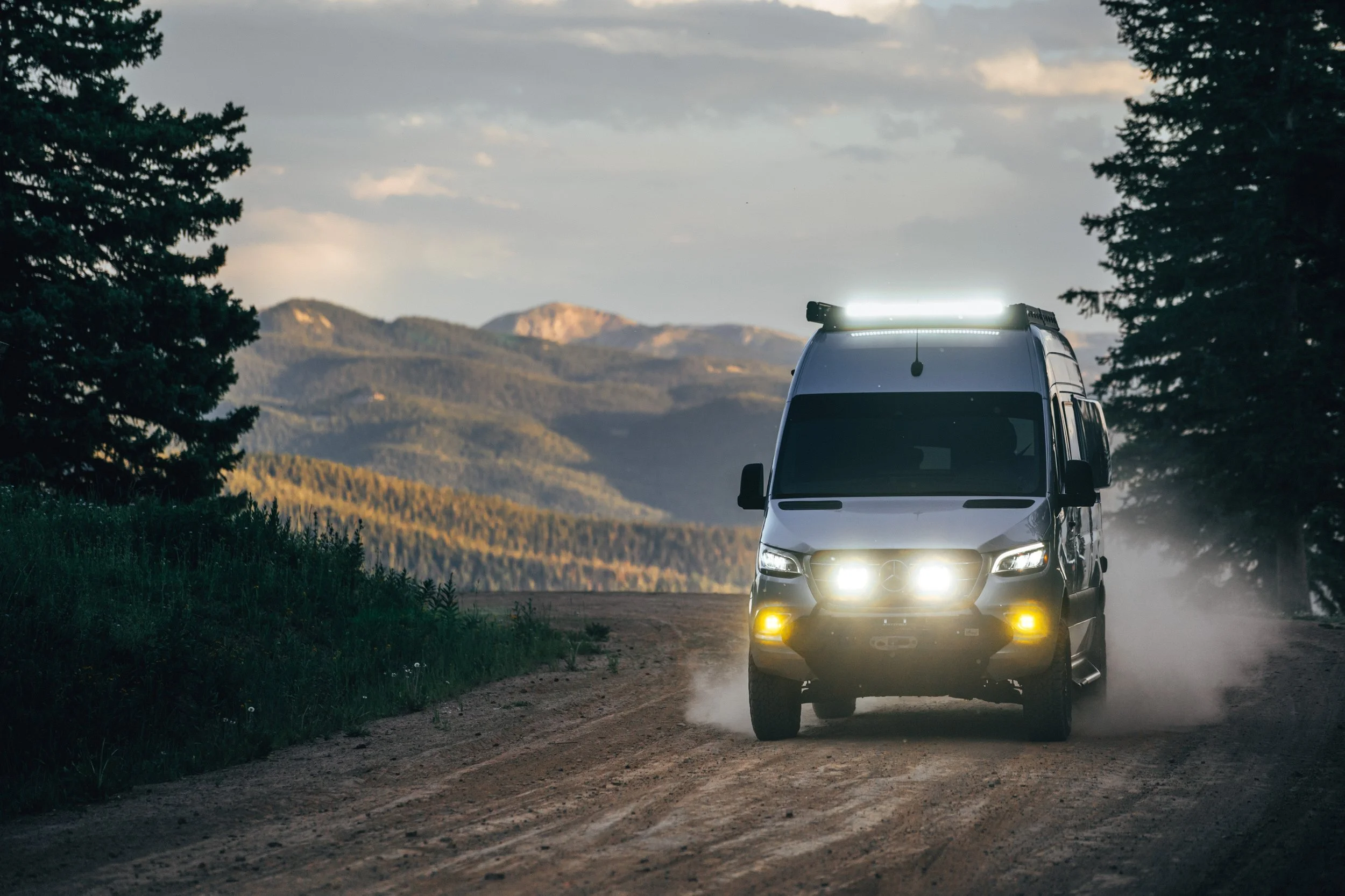 A black camper van driving on a dirt road in a mountainous landscape during dusk, with dust clouds behind it and trees on either side.