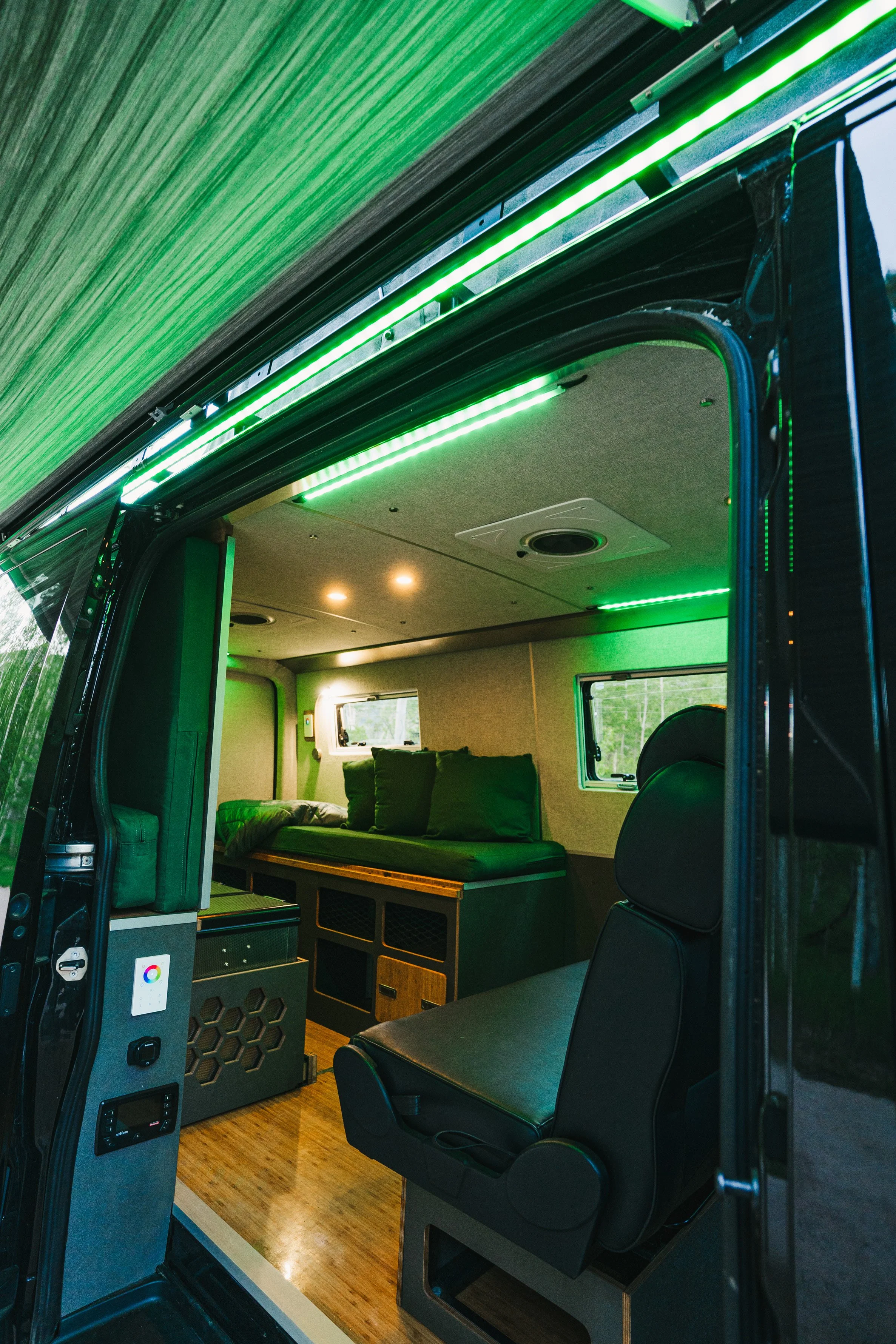 Inside a van converted into a camper or tiny home with green LED lighting, a sitting area with a padded bench, two windows, and a wooden floor.