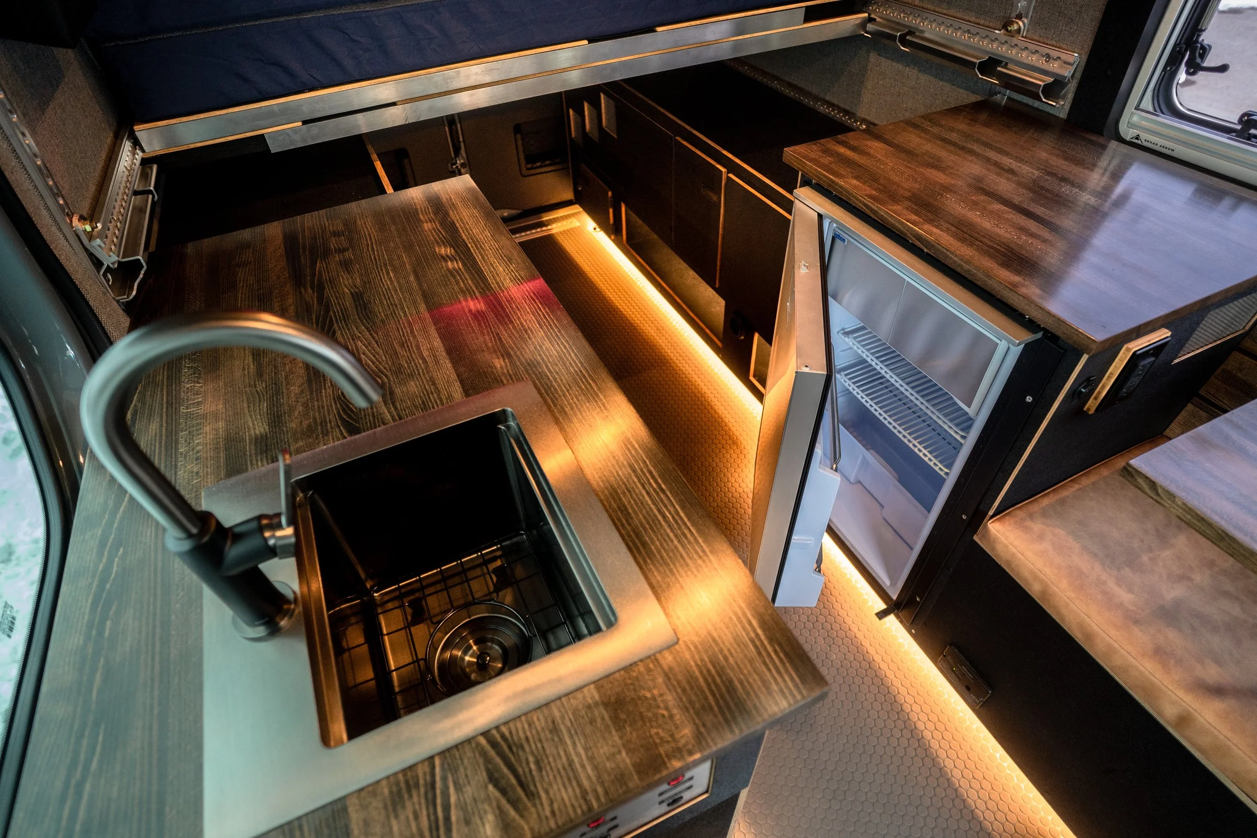 Interior of a compact kitchen with a wooden countertop, small sink with faucet, mini refrigerator, and storage cabinets, illuminated with warm under-cabinet lighting.
