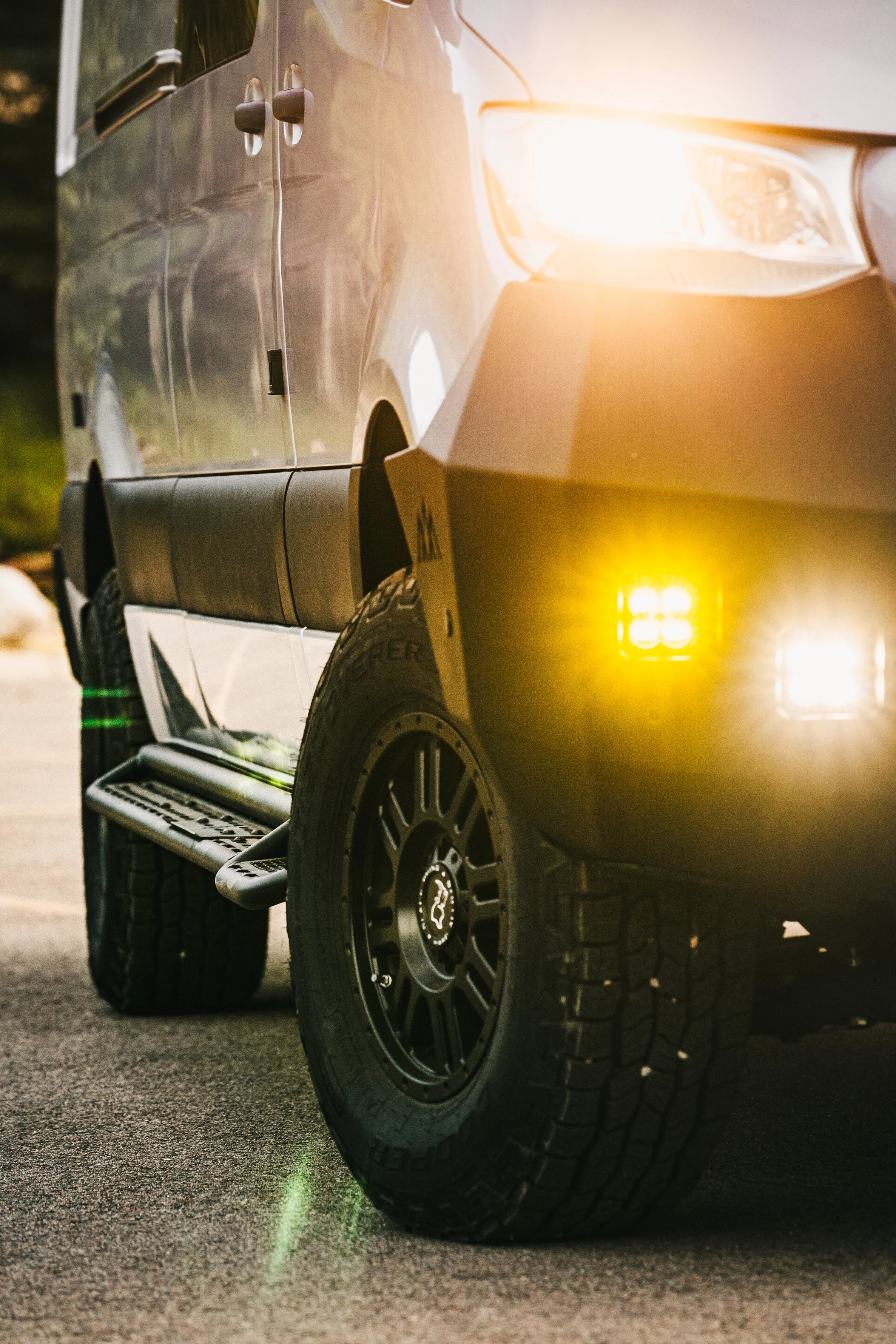 Close-up of a silver Sprinter van with yellow fog lights and a bright headlight, parked on a paved surface during sunset.