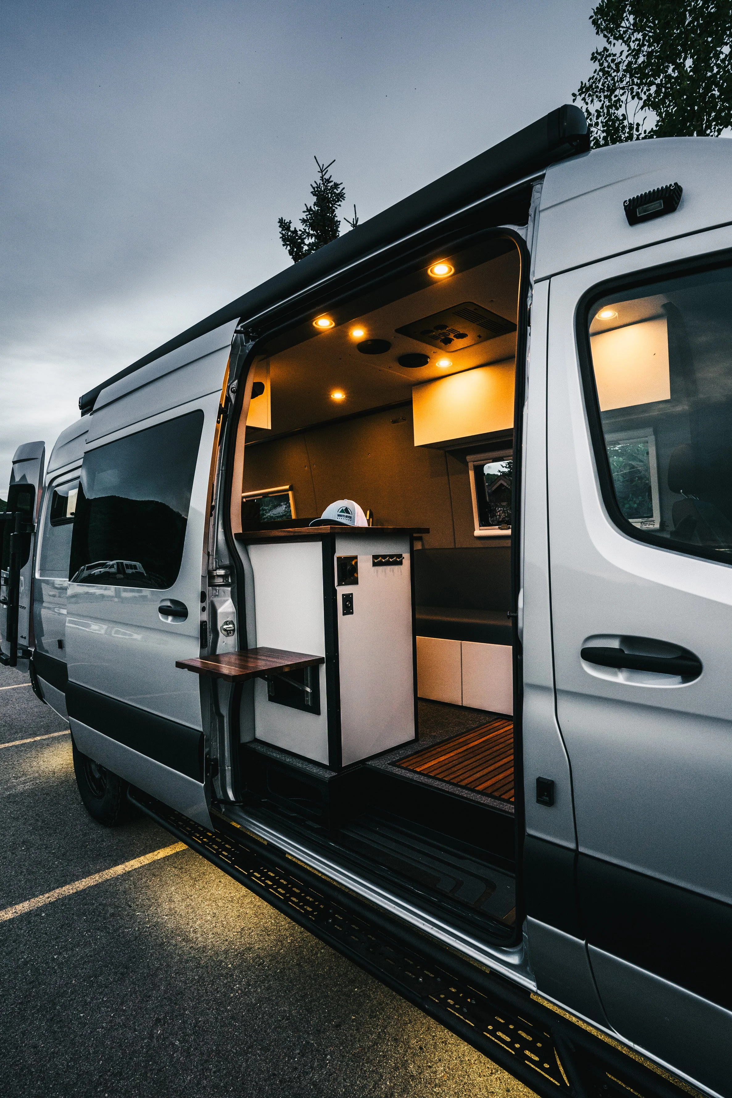 Inside view of a converted camper Sprinter van with lighting, kitchen counter, window, bench seat, and steps leading inside, parked on a paved lot with trees in the background.