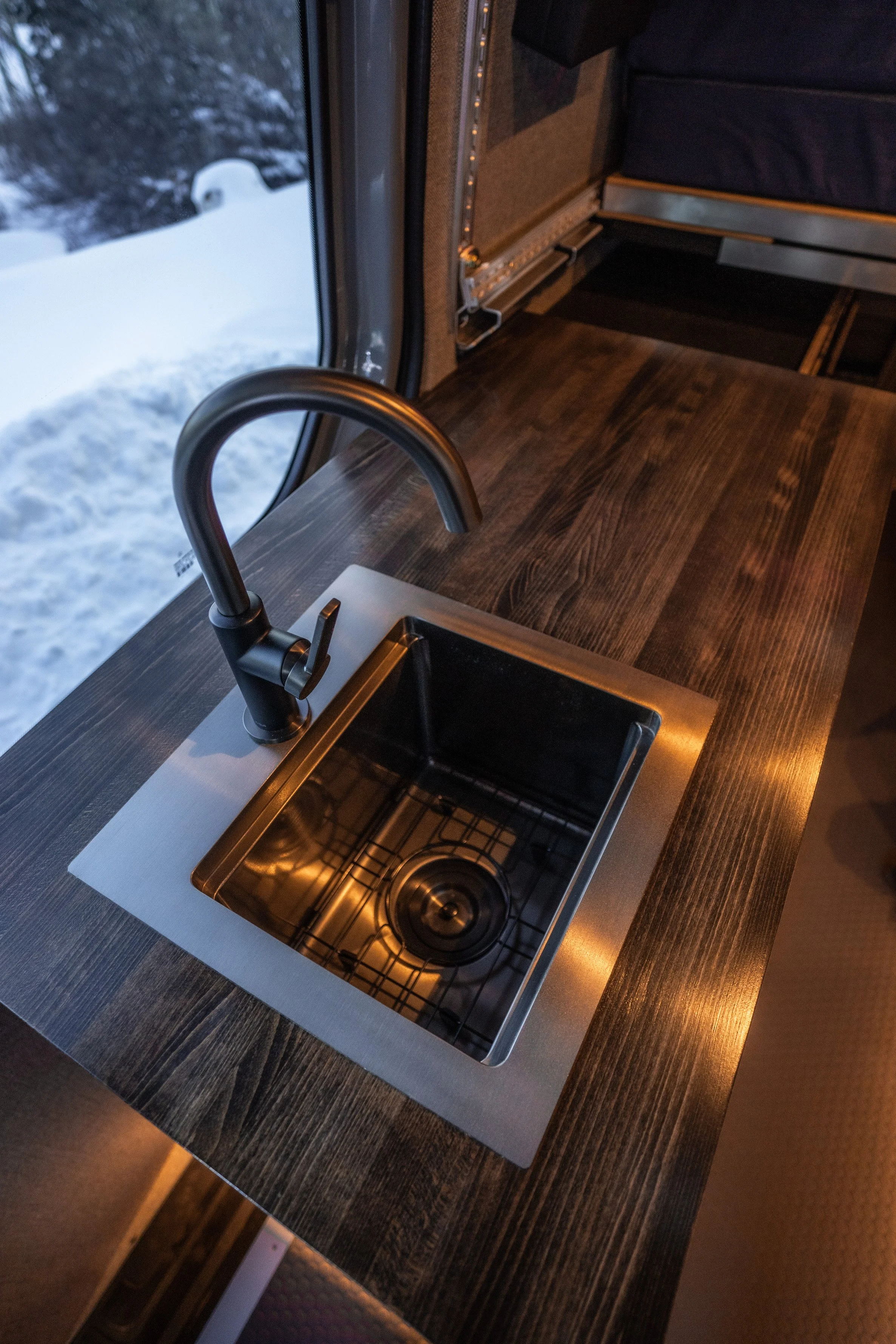 Interior of a vehicle with a wooden countertop, stainless steel sink, and a black faucet, next to a window showing snow outside.