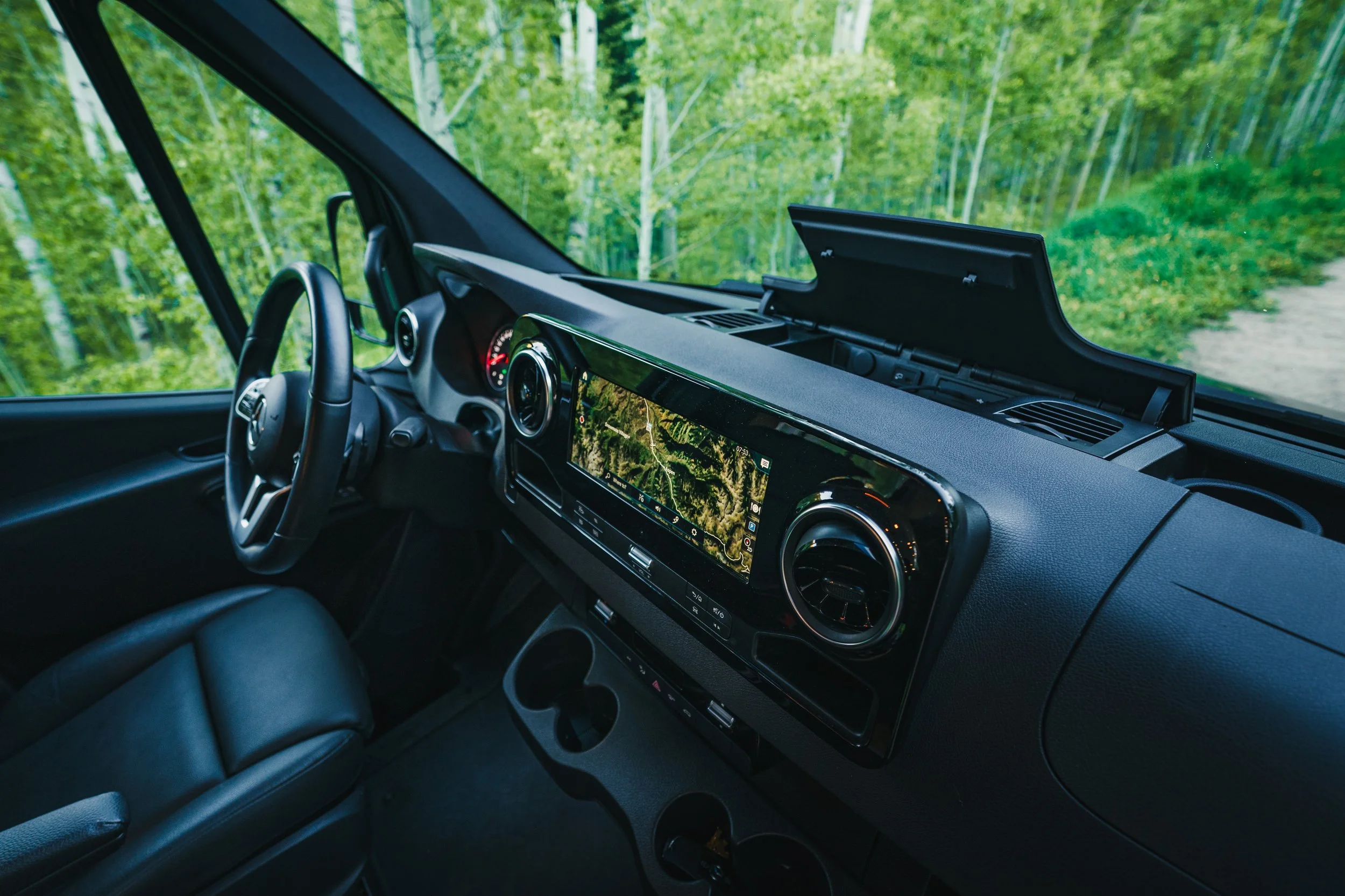 Interior of a vehicle showing a GPS navigation screen on the dashboard, steering wheel, and green forest scenery outside the windshield.